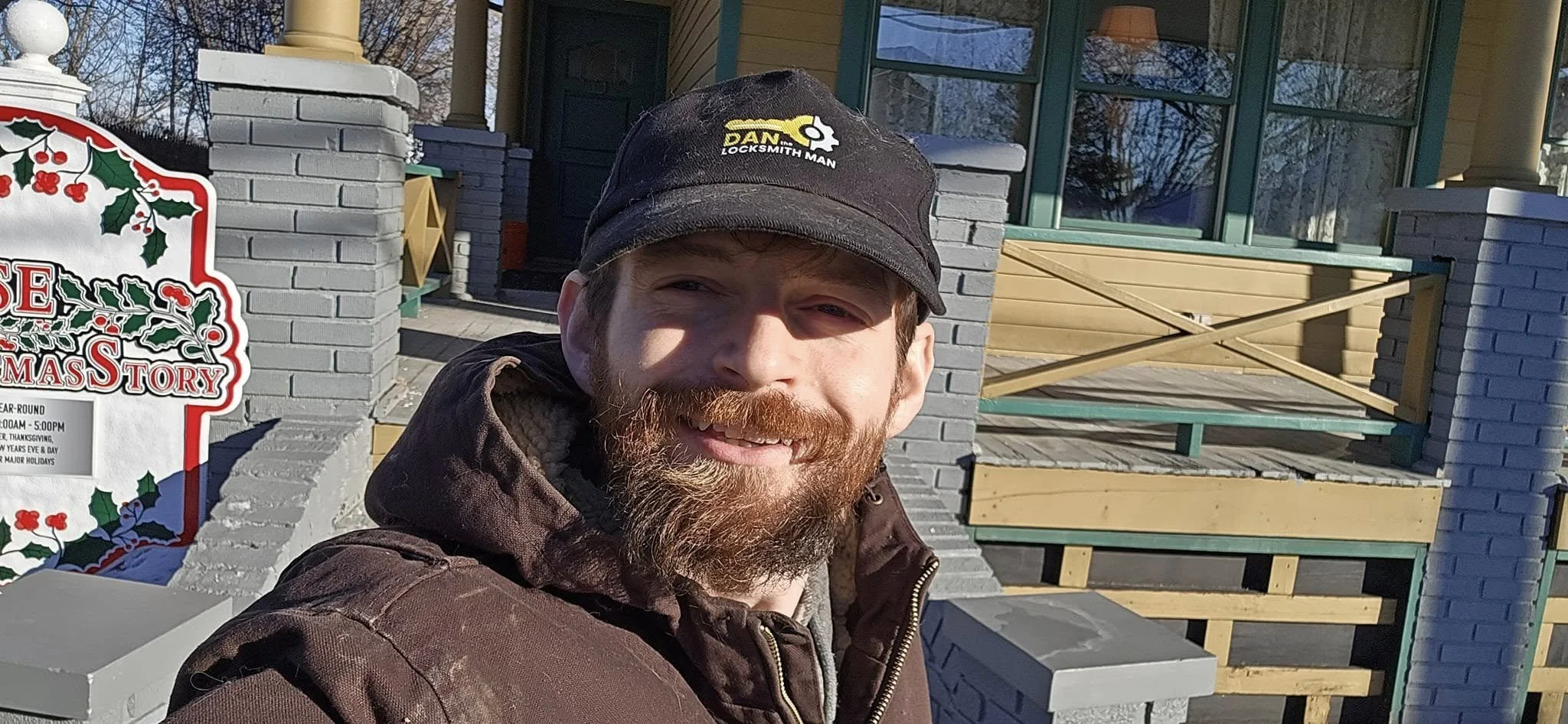 A man with a beard and mustache wearing a black cap with a locksmith logo, taking a selfie in front of a house porch with holiday wreaths and signs, during daytime with clear weather.