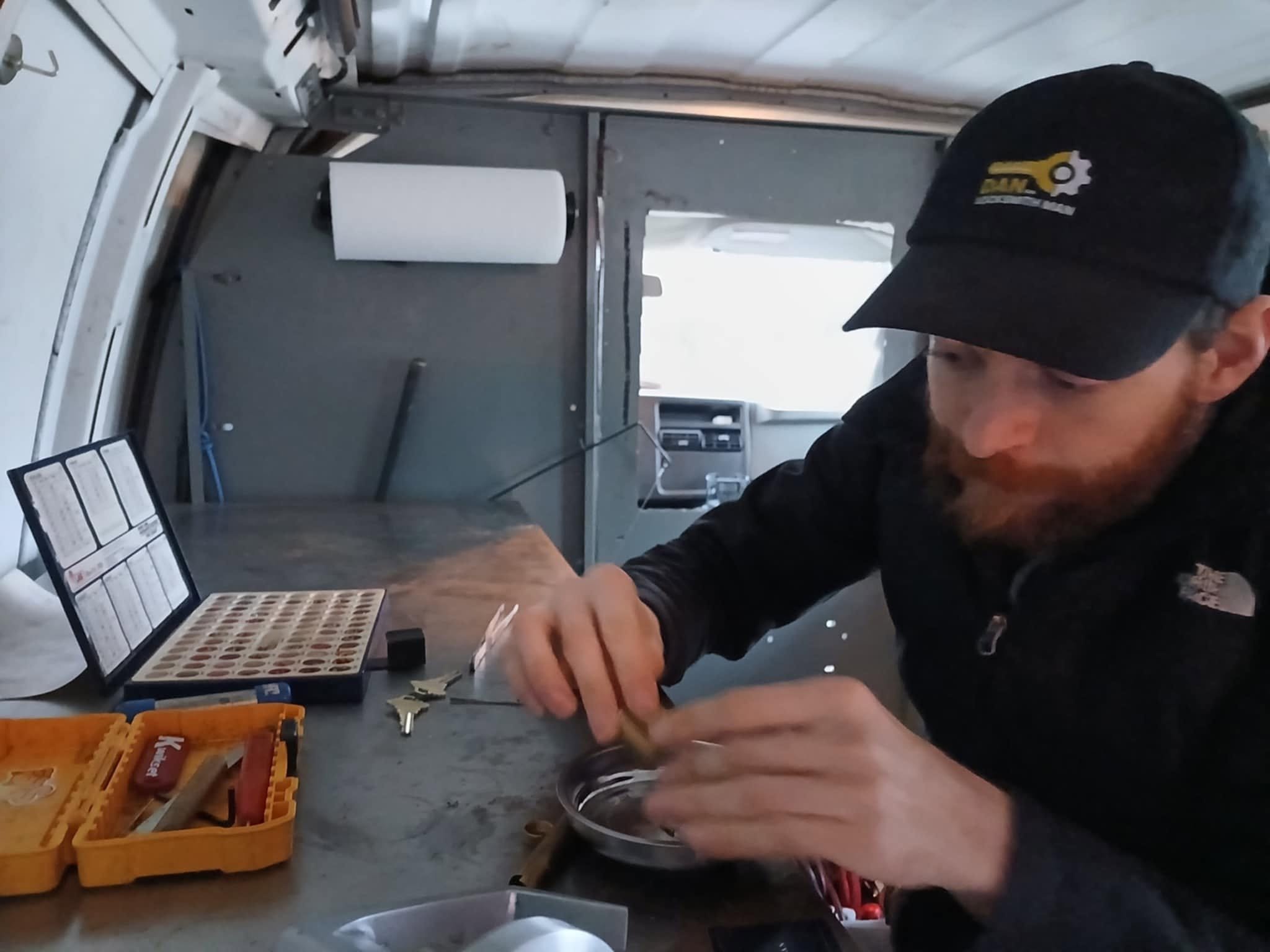 A man with a beard and dark hair wearing a black cap and black shirt working at a workbench. The workbench has a small black container, a yellow toolbox, and some small parts. The man appears to be assembling or repairing a small object. The background includes a metal door, a gray wall, and some kitchen appliances.