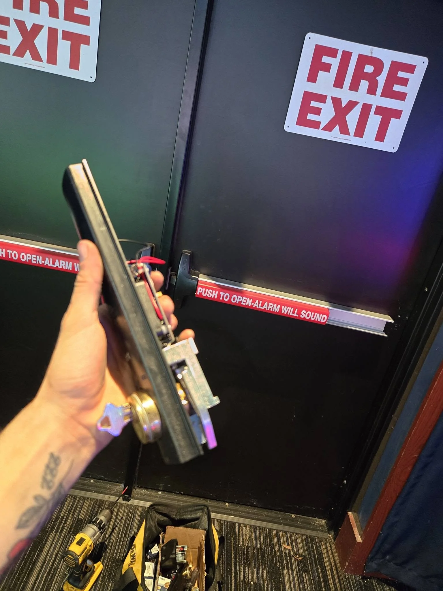 Black door with red and white Fire Exit signs and a fire alarm pull station at bottom. A person is holding a remote with a red string attached. The floor has a black and multicolored striped carpet, and there are tools and a toolbox on the ground.