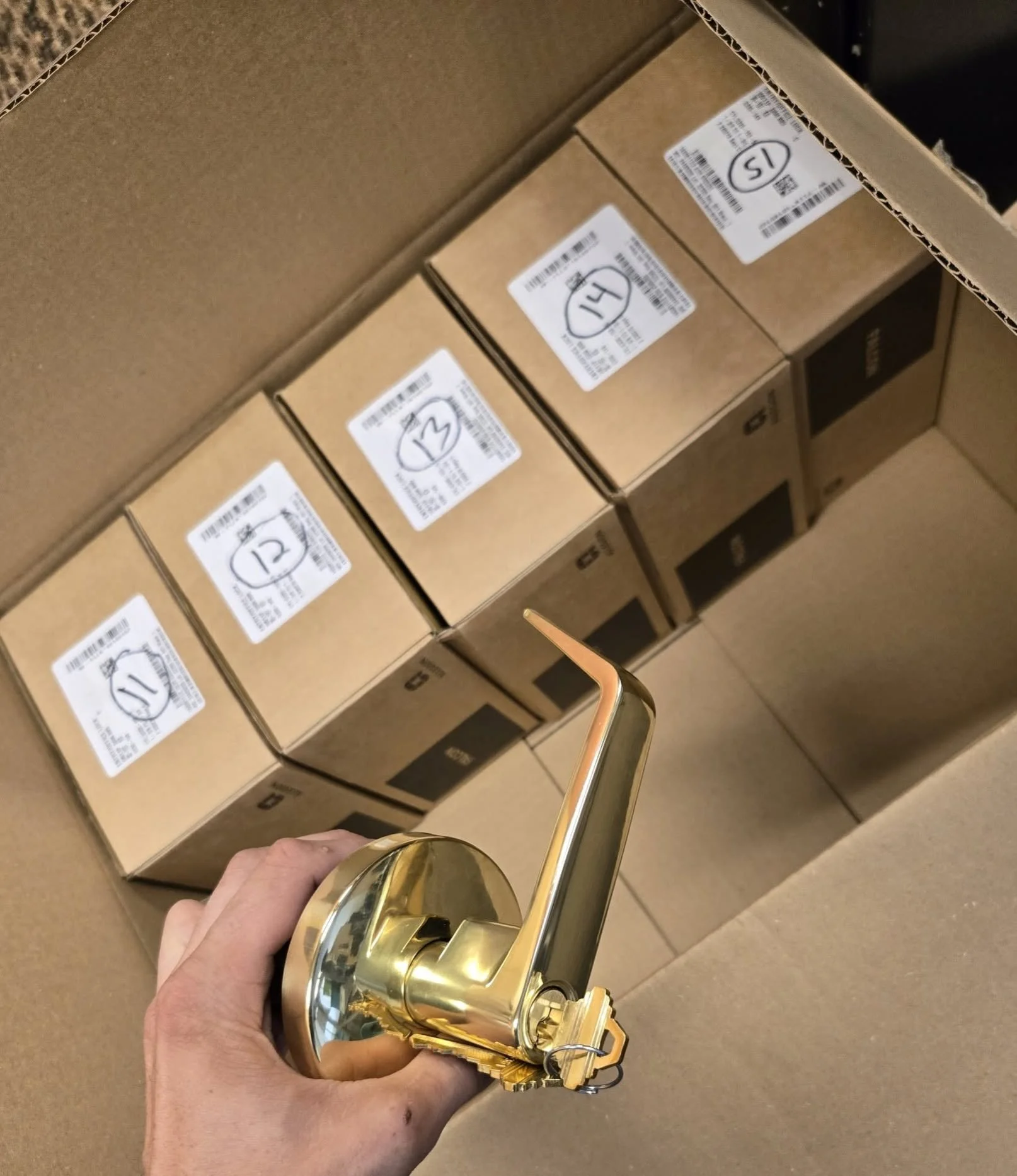 Hand holding a shiny gold door handle in front of stacked cardboard boxes with labels and numbers.