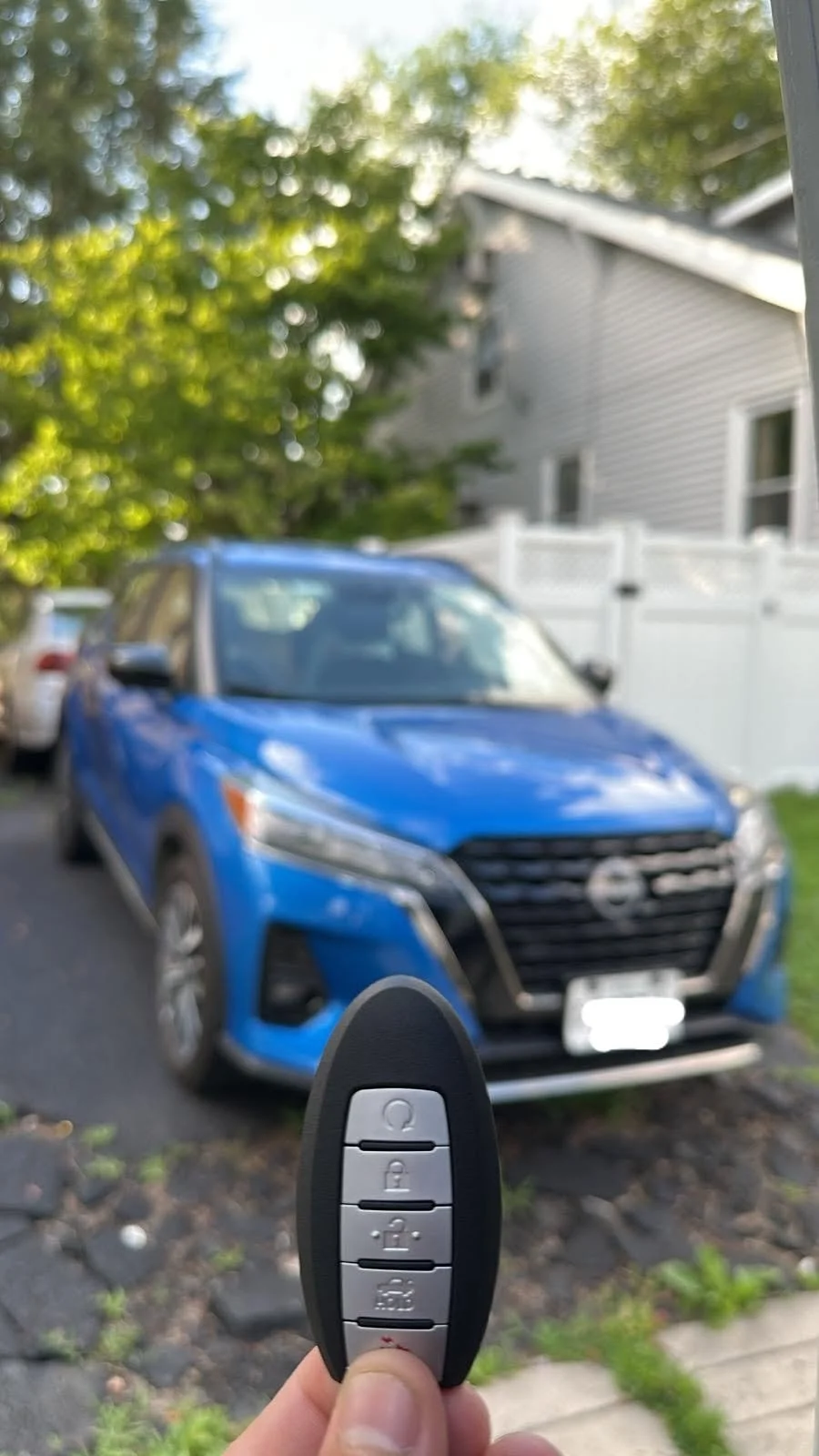 A person holding a car key fob in front of a blue car parked in a driveway, with a house and trees in the background.