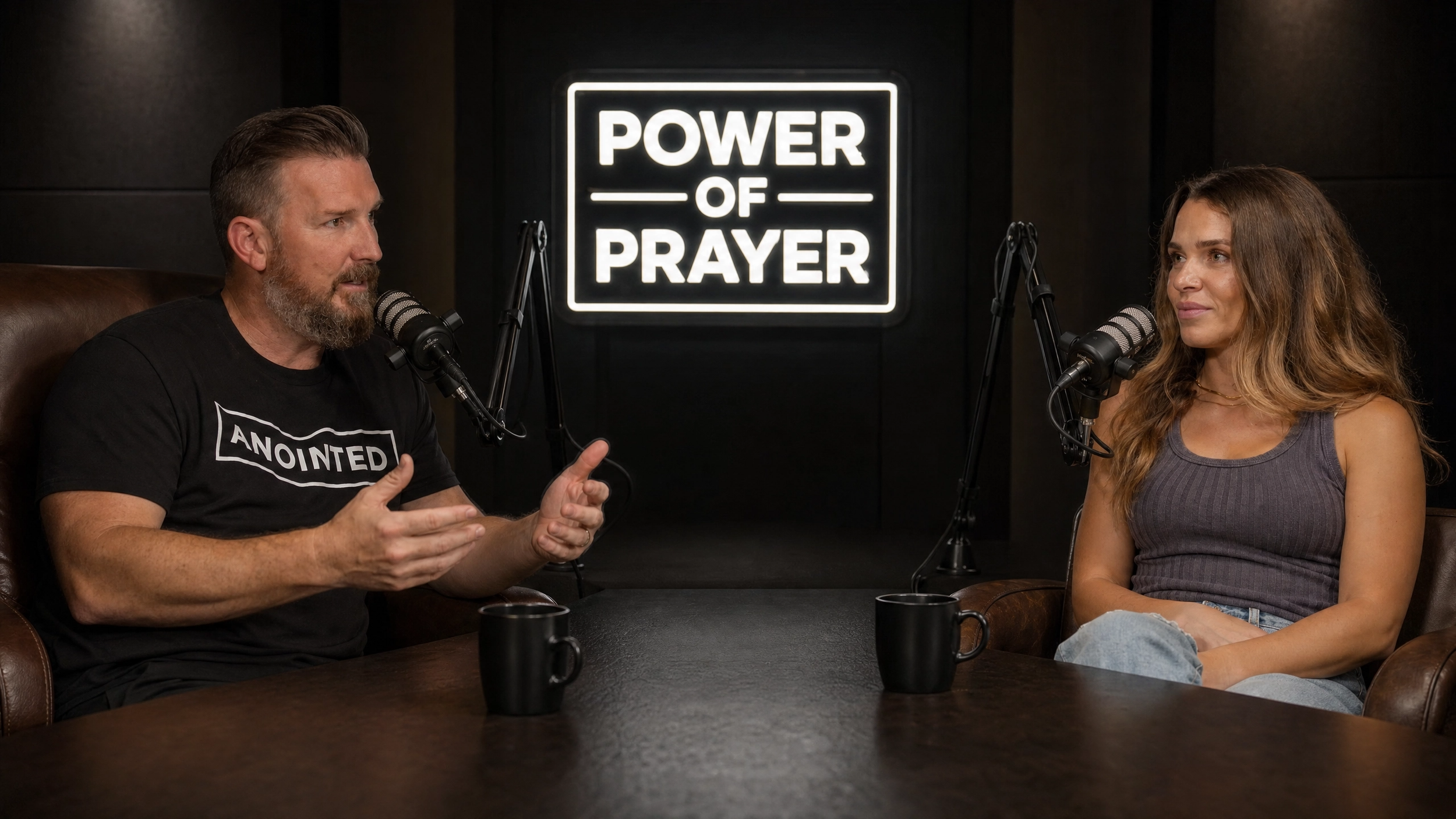 Two people, a man and a woman, speaking into microphones during a podcast recording with a sign behind them that reads 'Power of Prayer'.