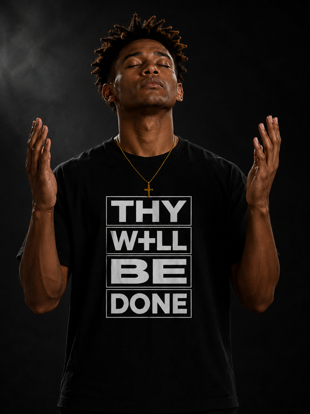 A young man with eyes closed and hands raised, wearing a black T-shirt with the words 'THY WILL BE DONE' in white within boxes, and a gold cross necklace, standing against a dark background.