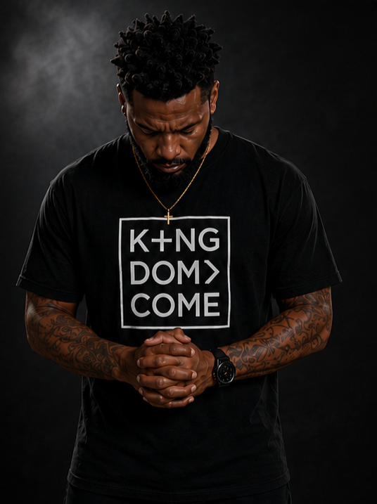 Man with tattoos on arms, wearing a black t-shirt with the words 'KINGDOM COME' inside a box and a cross necklace, praying or reflecting with hands clasped, dark background.