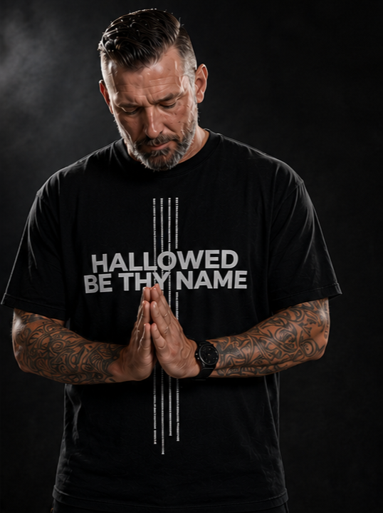 Man praying with hands pressed together, wearing a black T-shirt that says "Hallowed Be Thy Name," against a dark background.