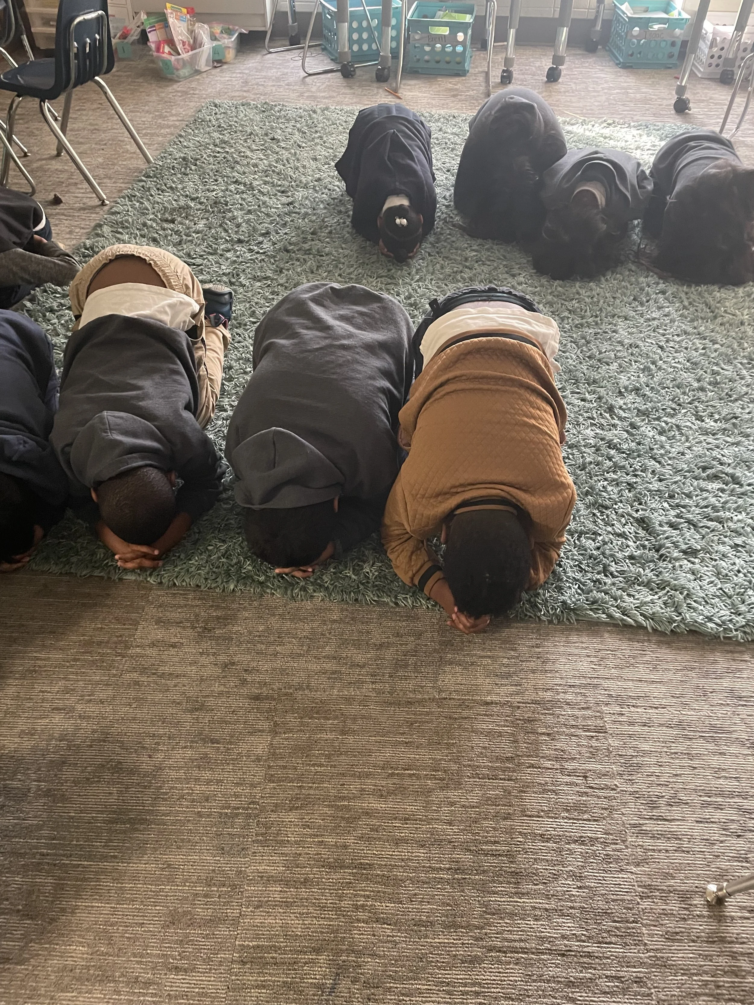 Six children in a classroom are kneeling on a green rug, with their hands clasped and heads bowed, possibly praying.