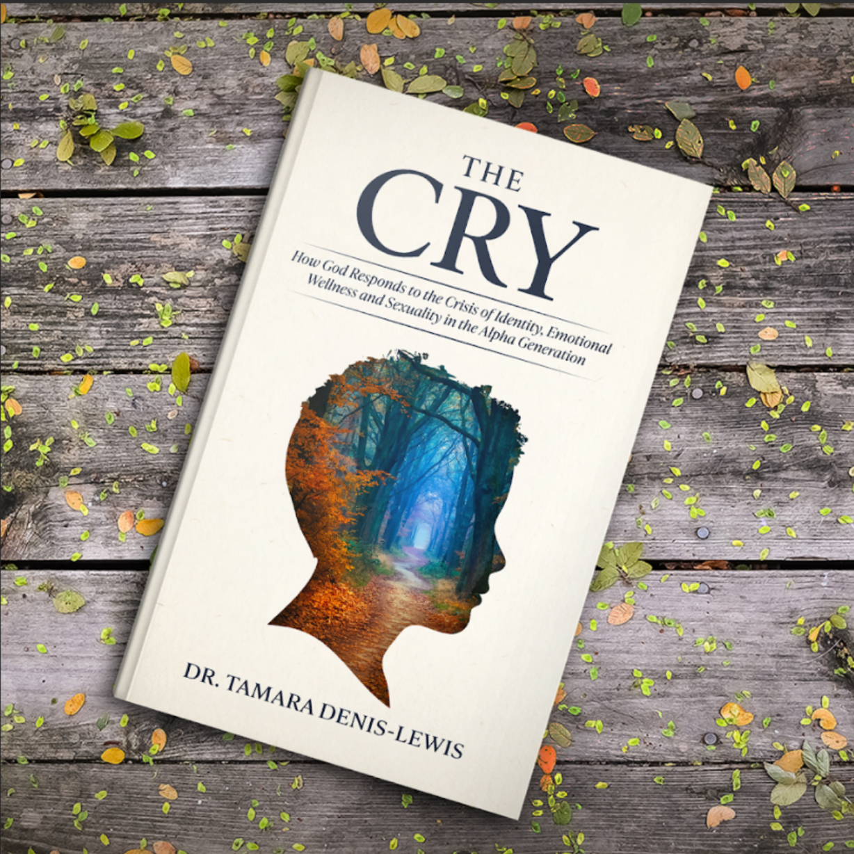 Book titled "The Cry" by Dr. Tamara Denis-Lewis, featuring a forest scene inside a silhouette of a person's profile, placed on a wooden surface with scattered leaves.