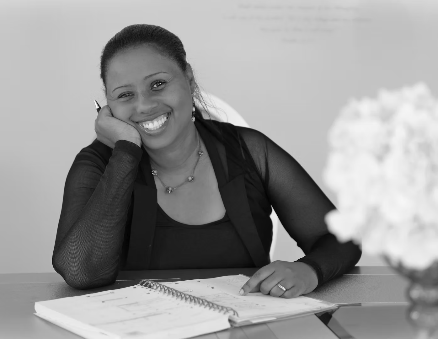 A smiling woman with a notebook open in front of her, sitting at a desk with a pen in her hand, in an office setting.