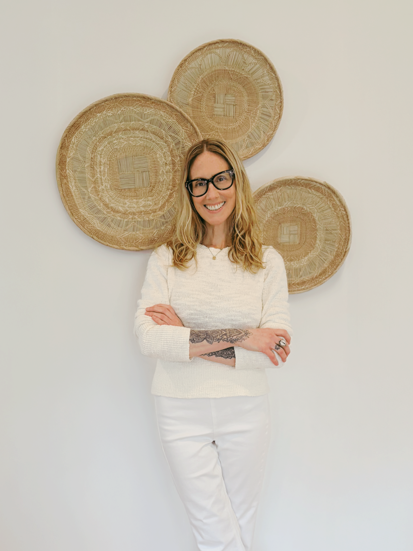 Kelly Chapman, LCSW, smiling with arms crossed in front of woven wall baskets.