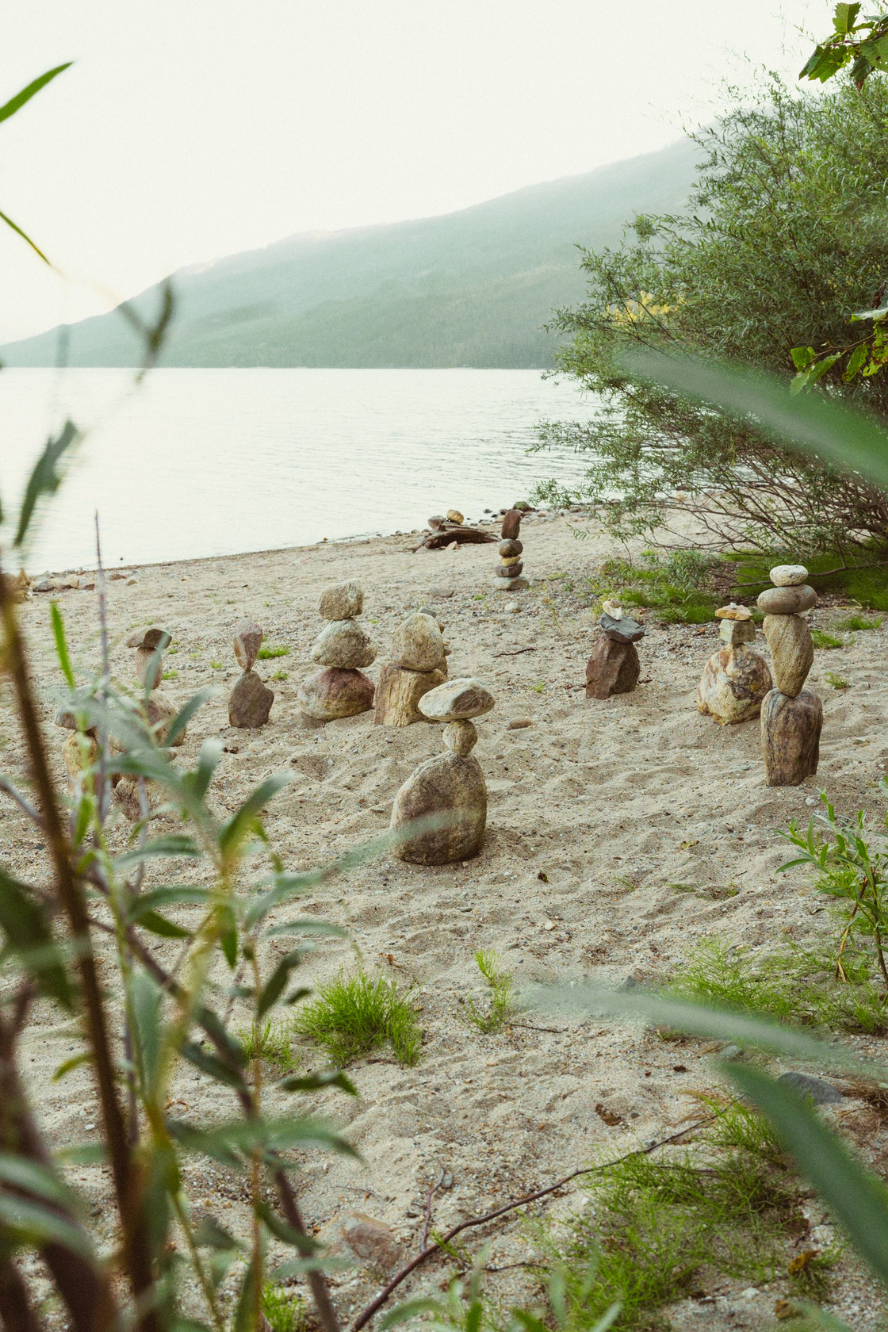 Balanced stone cairns on a sandy lakeshore, with still water and a misty mountain behind.