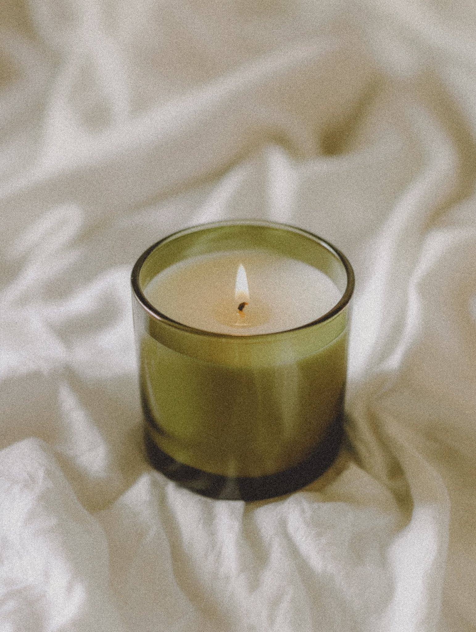 Lit scented candle in olive glass on cream textured fabric.