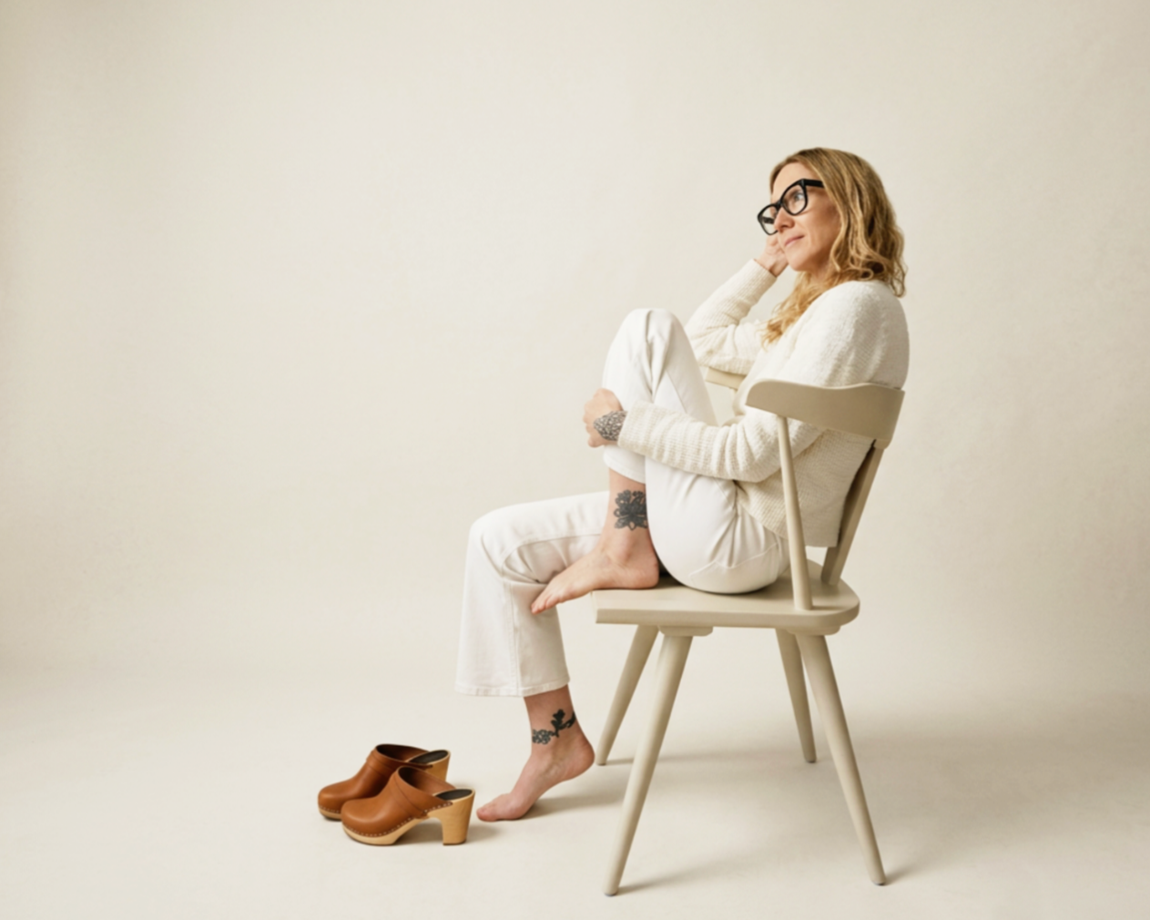 Kelly Chapman, LCSW, seated barefoot on a wooden chair with her knees drawn up, looking off to the side.