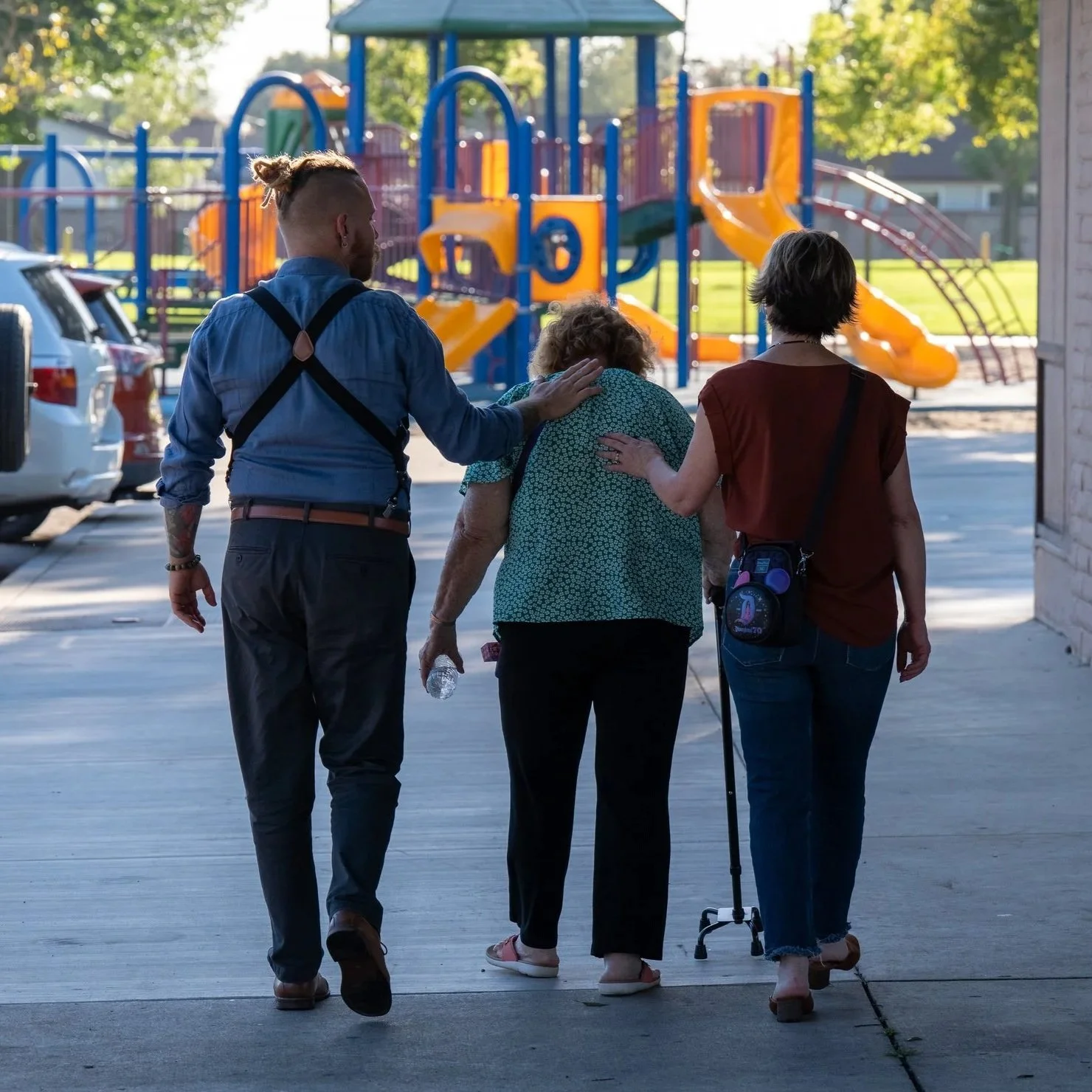 A group of three people walking away from the camera towards a colorful playground, with one elderly woman using a cane, accompanied by Brandon Schooner with his hand on her shoulder and a woman leading them.