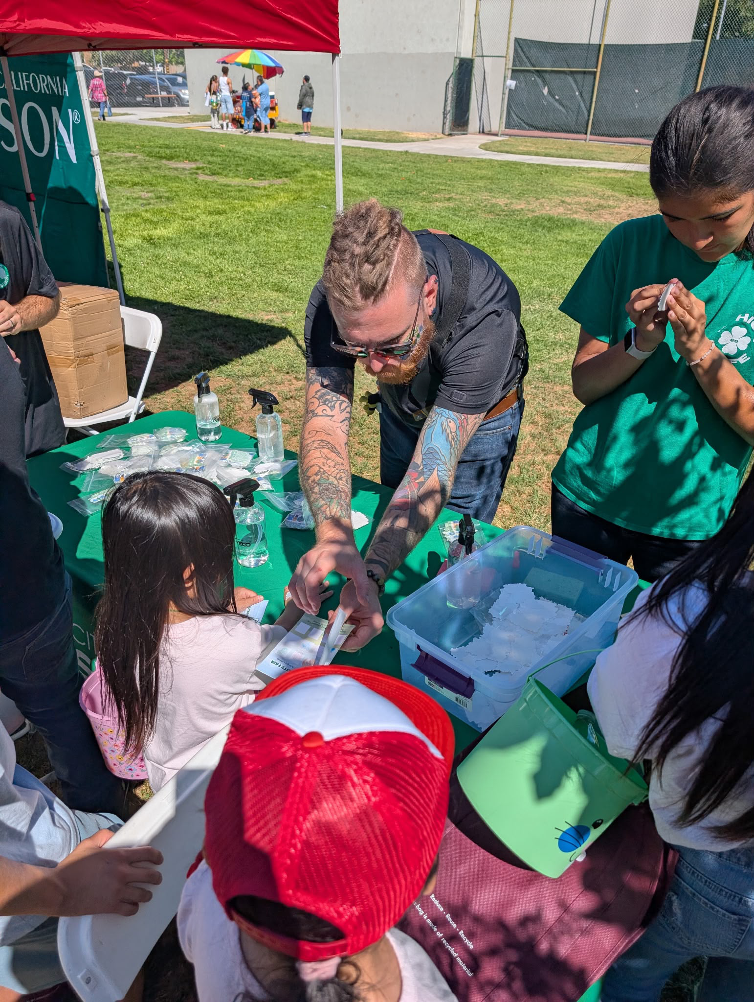 People gathered around a table outdoors at a community event, with Brandon Schooner handing out temporary tattoos ans stickers to children, a red tent and a colorful umbrella, with a sports field and parking lot visible in the background