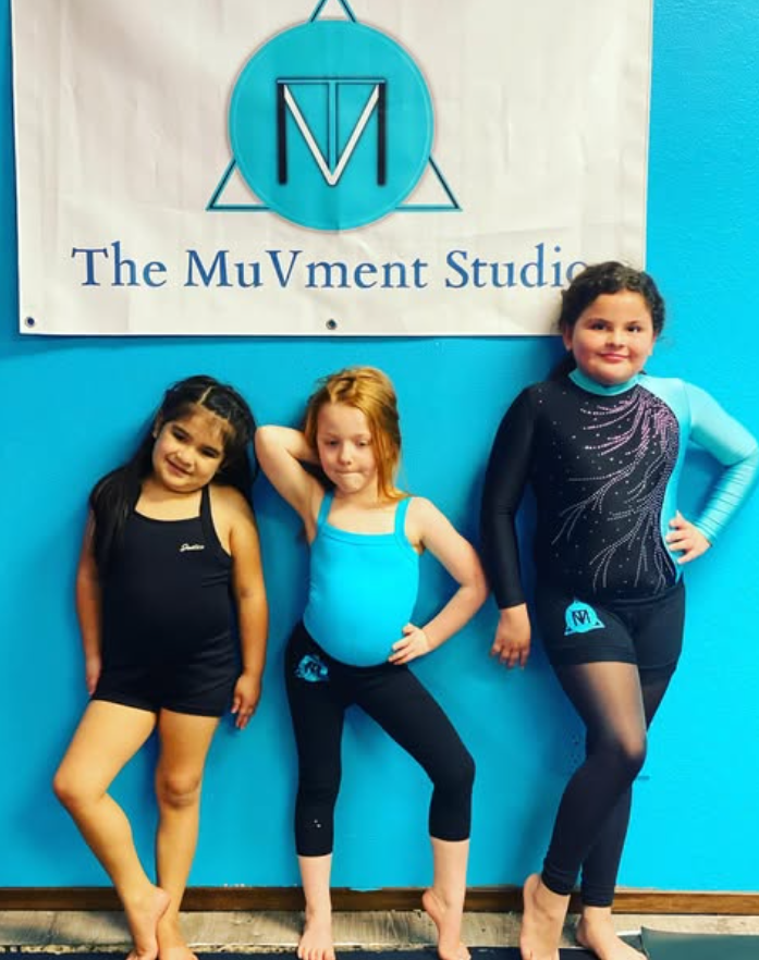 Three young girls in gymnastics outfits posing in front of a sign that reads "The MuVment Studio" with a blue and black logo above.