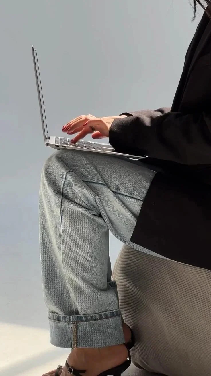 Person sitting on a beige cushioned chair, wearing a black blazer, gray rolled-up jeans, and black open-toe heels, typing on a silver laptop with a minimalistic gray background.