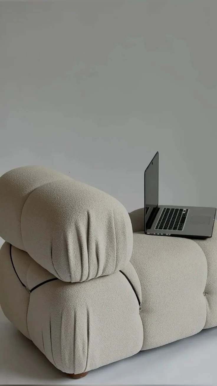 A beige, plush, modern lounge chair with a small, open laptop on a side extension.