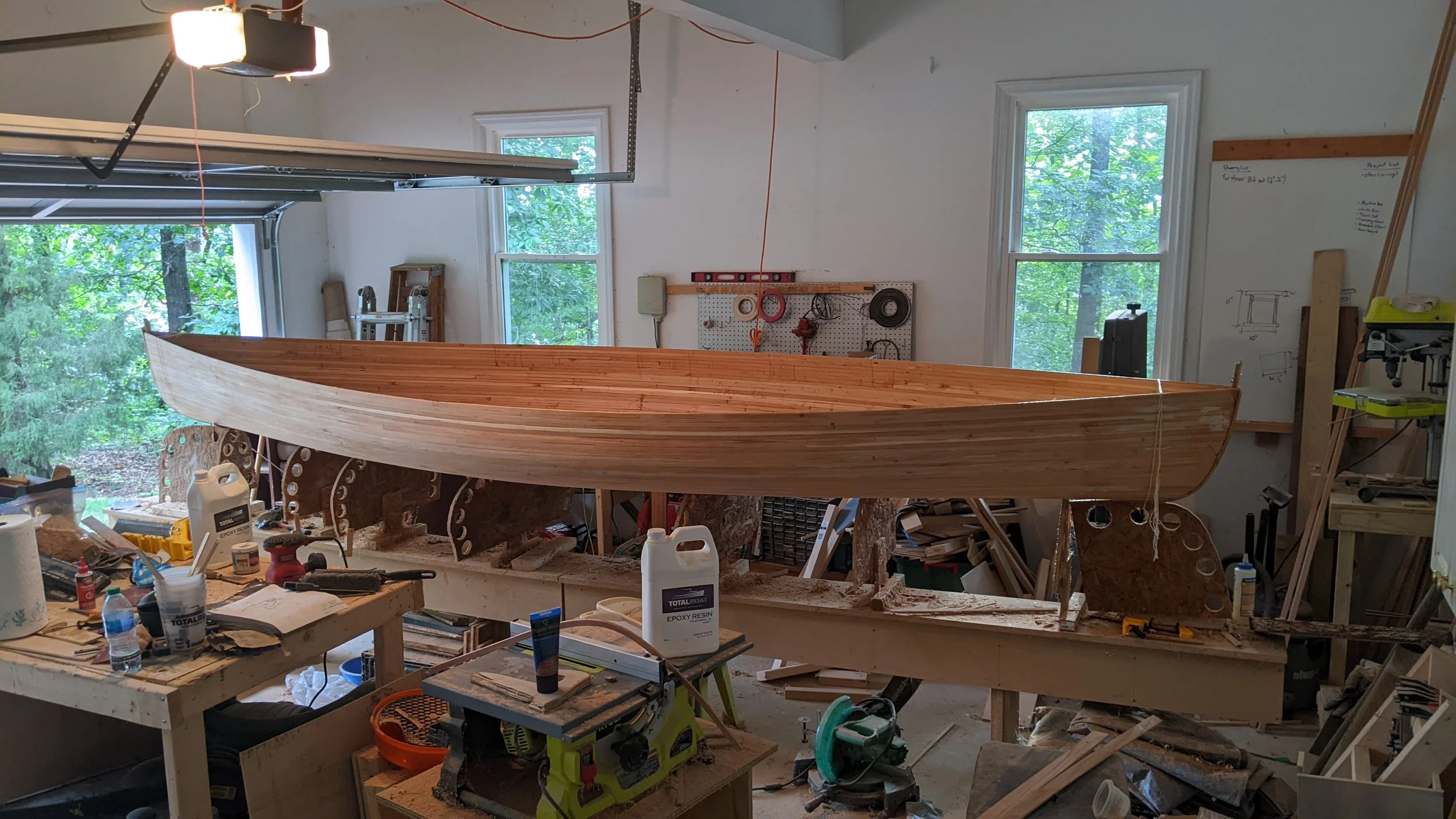 Wooden boat in construction in a workshop with tools, supplies, and workbenches around, windows with greenery outside in background.