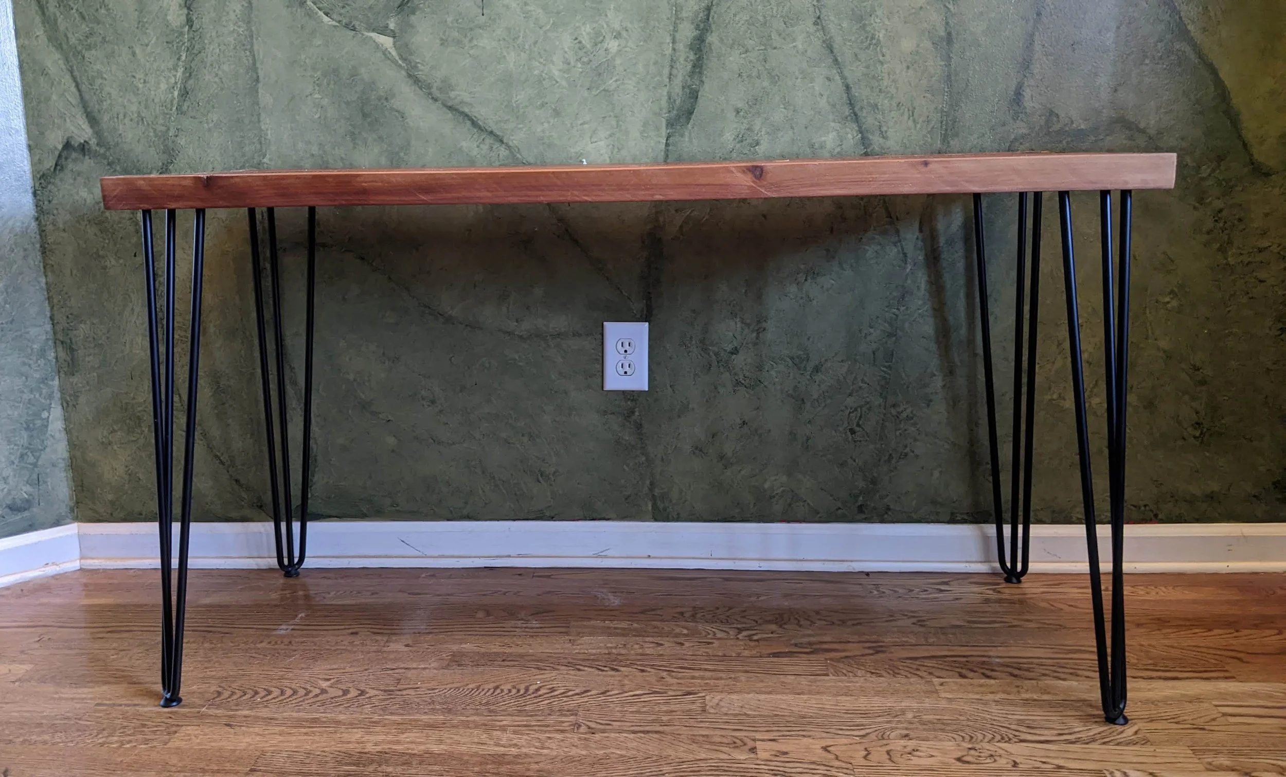 A wooden table with black metal hairpin legs against a green textured wall and a wooden floor.