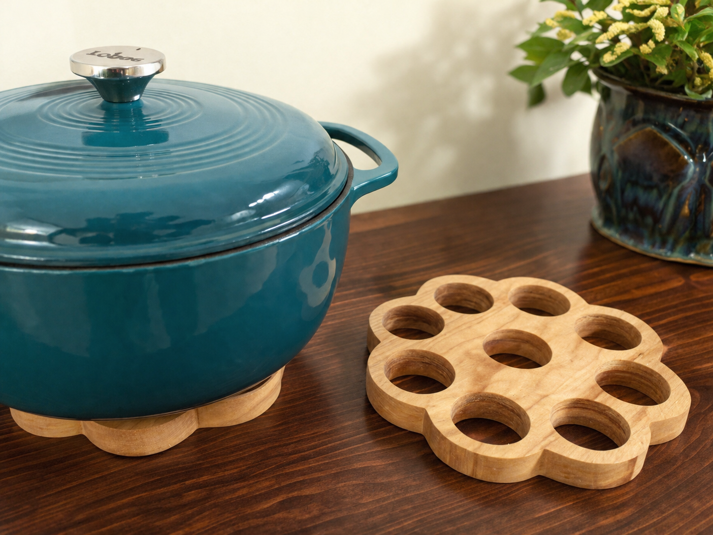 Blue enameled cast iron Dutch oven with a lid on a wooden trivet, a wooden trivet with circular holes, and a ceramic pot with flowers on a wooden table.
