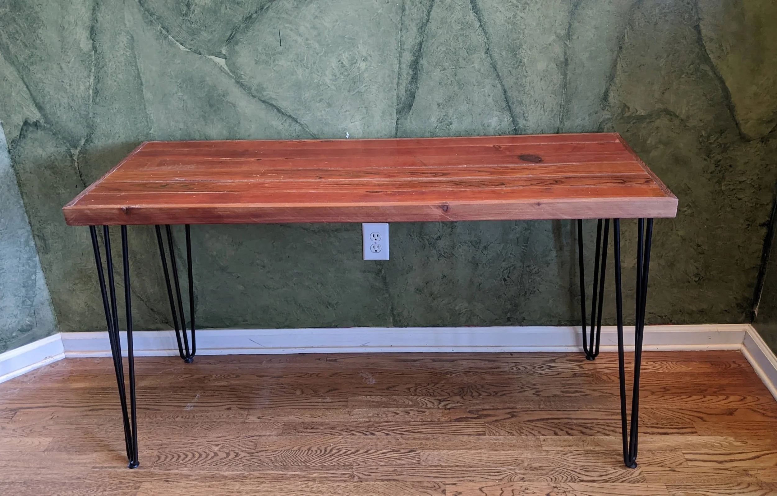 A wooden tabletop with a metal hairpin leg frame, placed against a green textured wall and a wood floor.