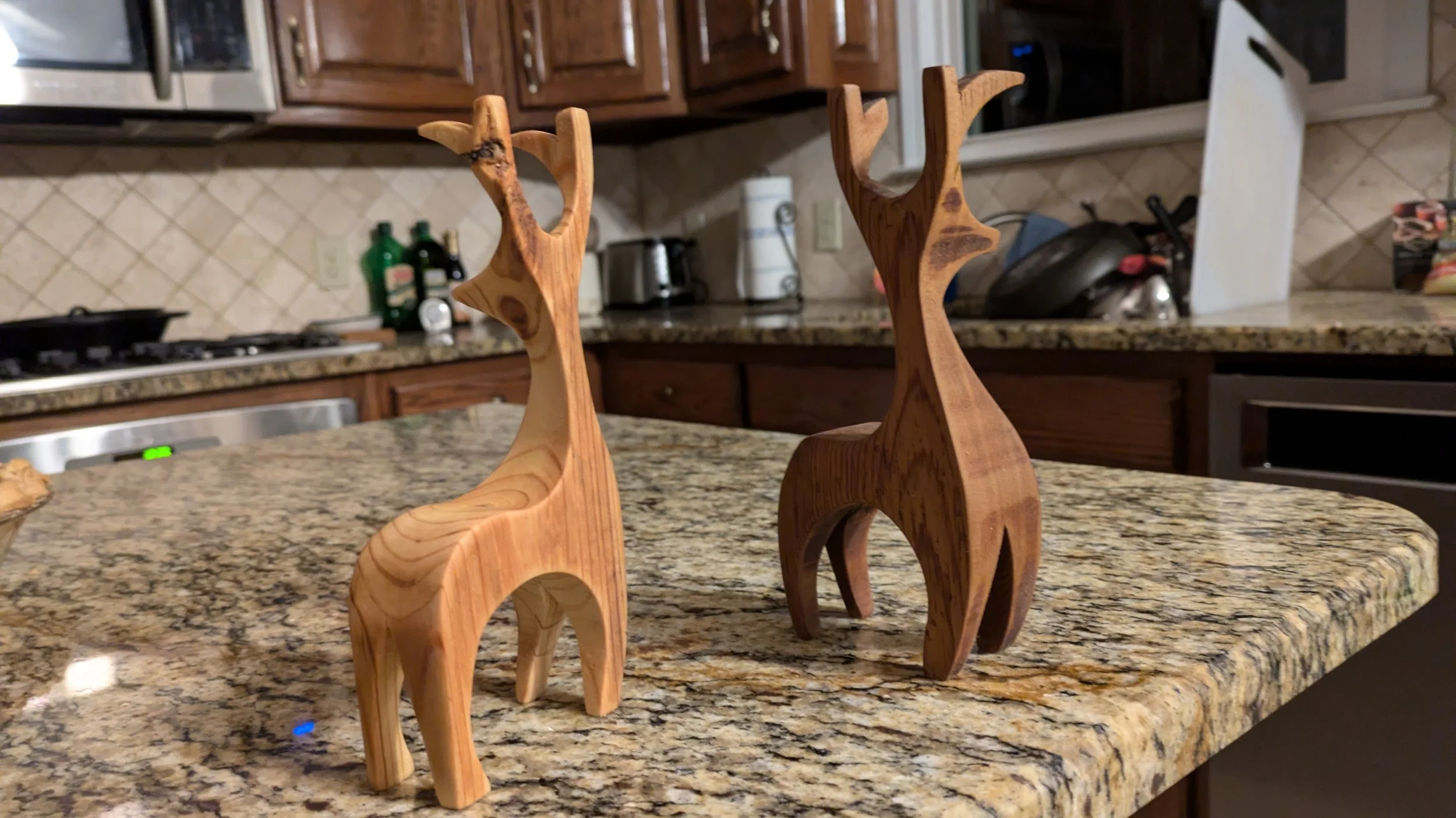 Two wooden reindeer figurines on a granite kitchen countertop with wooden cabinets, a stove, and various kitchen items in the background.