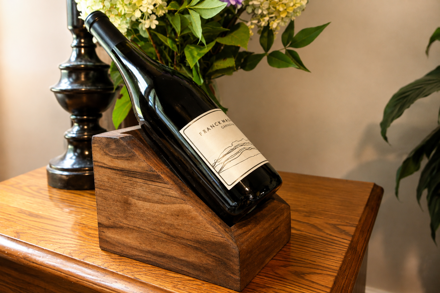 A wine bottle labeled 'FRANC MAC' resting on a wooden wine holder on a polished wooden table, with a black decorative lamp and green plants in the background.