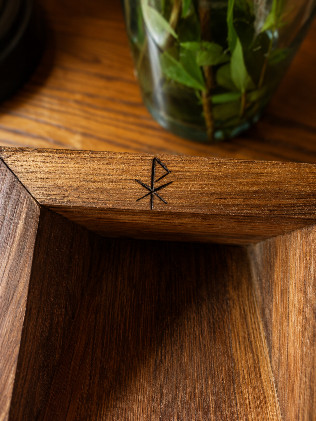 Close-up of a wooden surface with a decorative carved star pattern, with a glass vase with green leaves in the background.