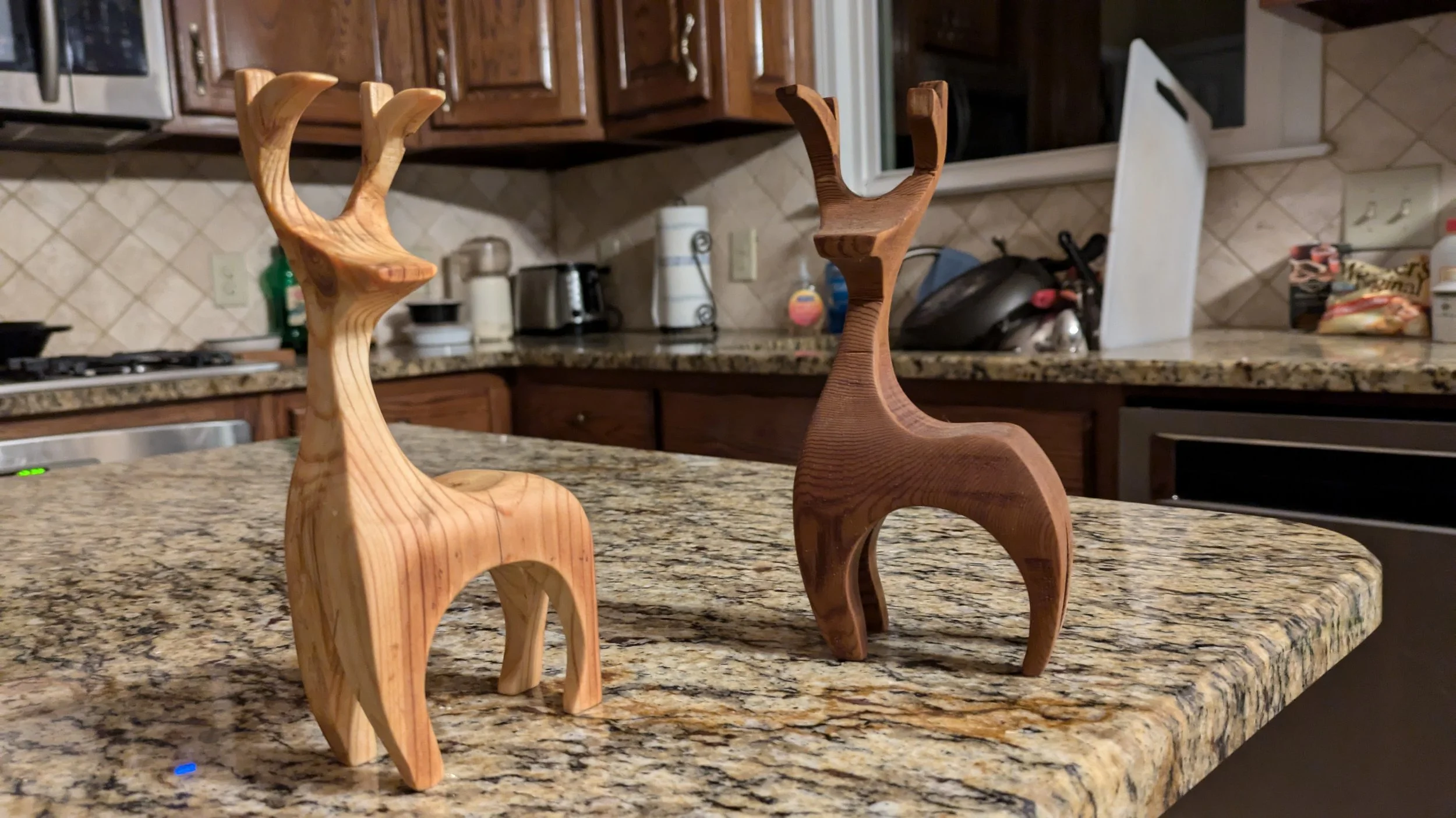 Two wooden reindeer sculptures on a kitchen countertop with brown and beige granite. The kitchen has dark wooden cabinets, a stove, and various appliances and items in the background.