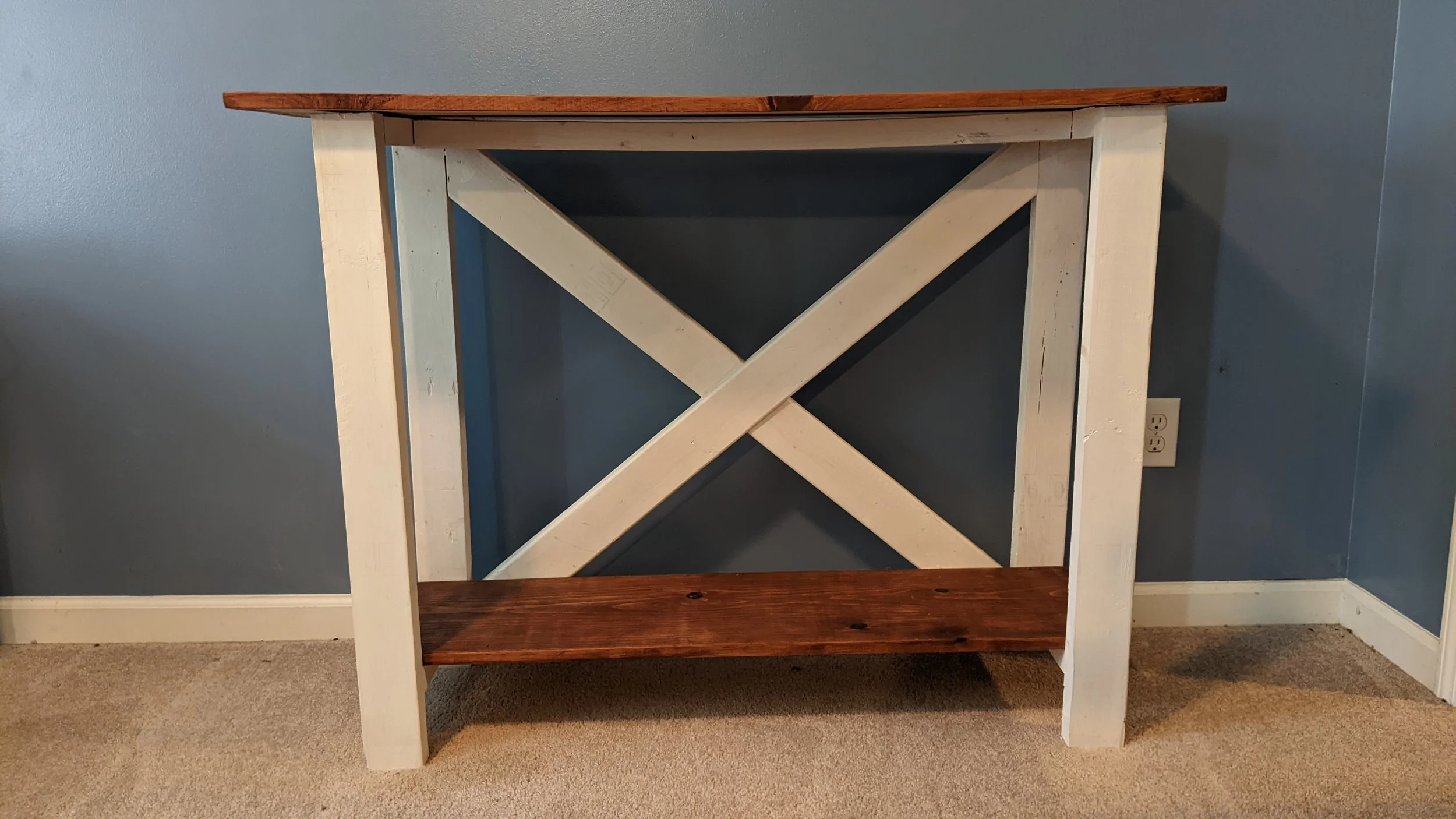 A wooden table with a natural wood top and white painted legs and supports, placed against a blue wall with a white electrical outlet visible near the floor.