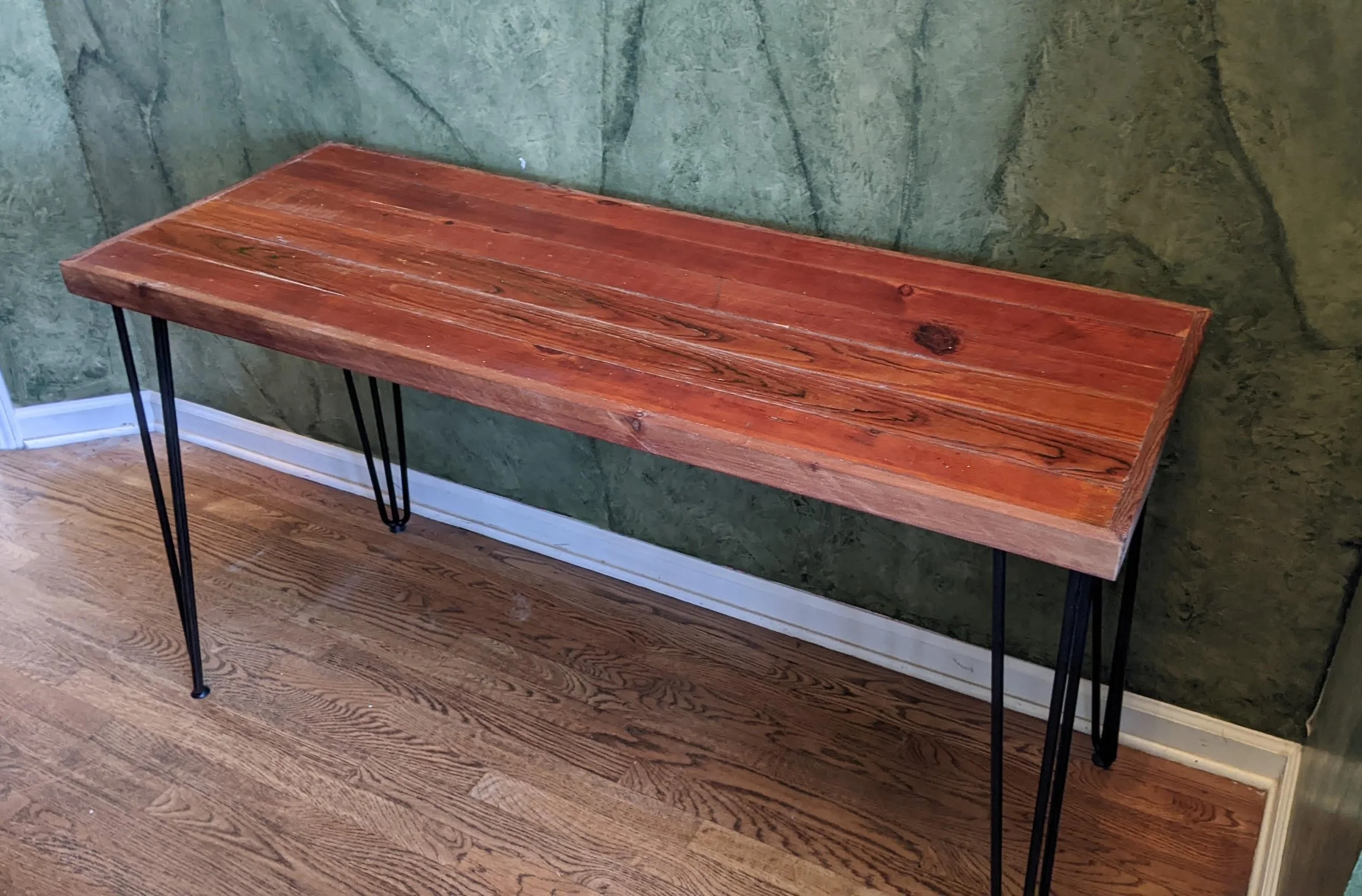 A wooden table with black metal hairpin legs is positioned against a green marble wall, with hardwood flooring below.