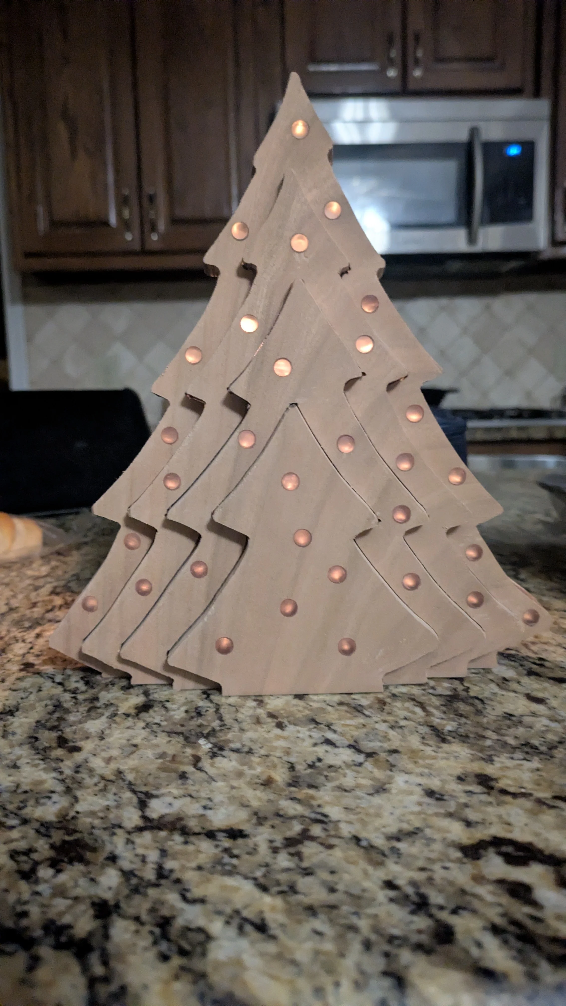 A decorative wooden Christmas tree with illuminated holes, placed on a kitchen countertop with a granite surface. In the background, there are dark wooden cabinets and a stainless steel microwave oven.