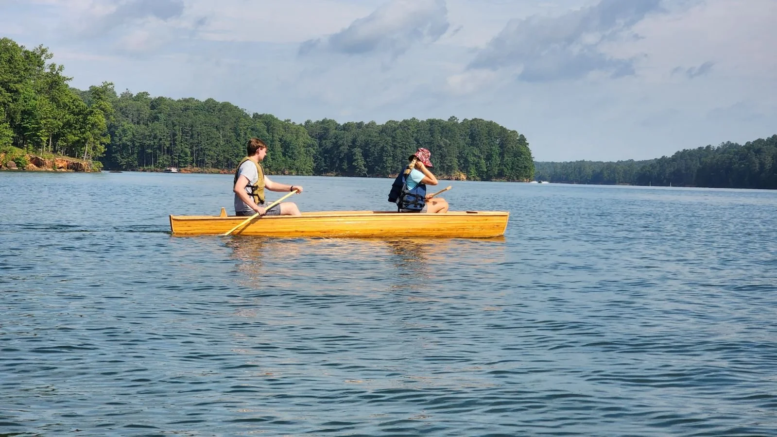 Two people paddling a small wooden boat on a large lake with forested shoreline in the background.