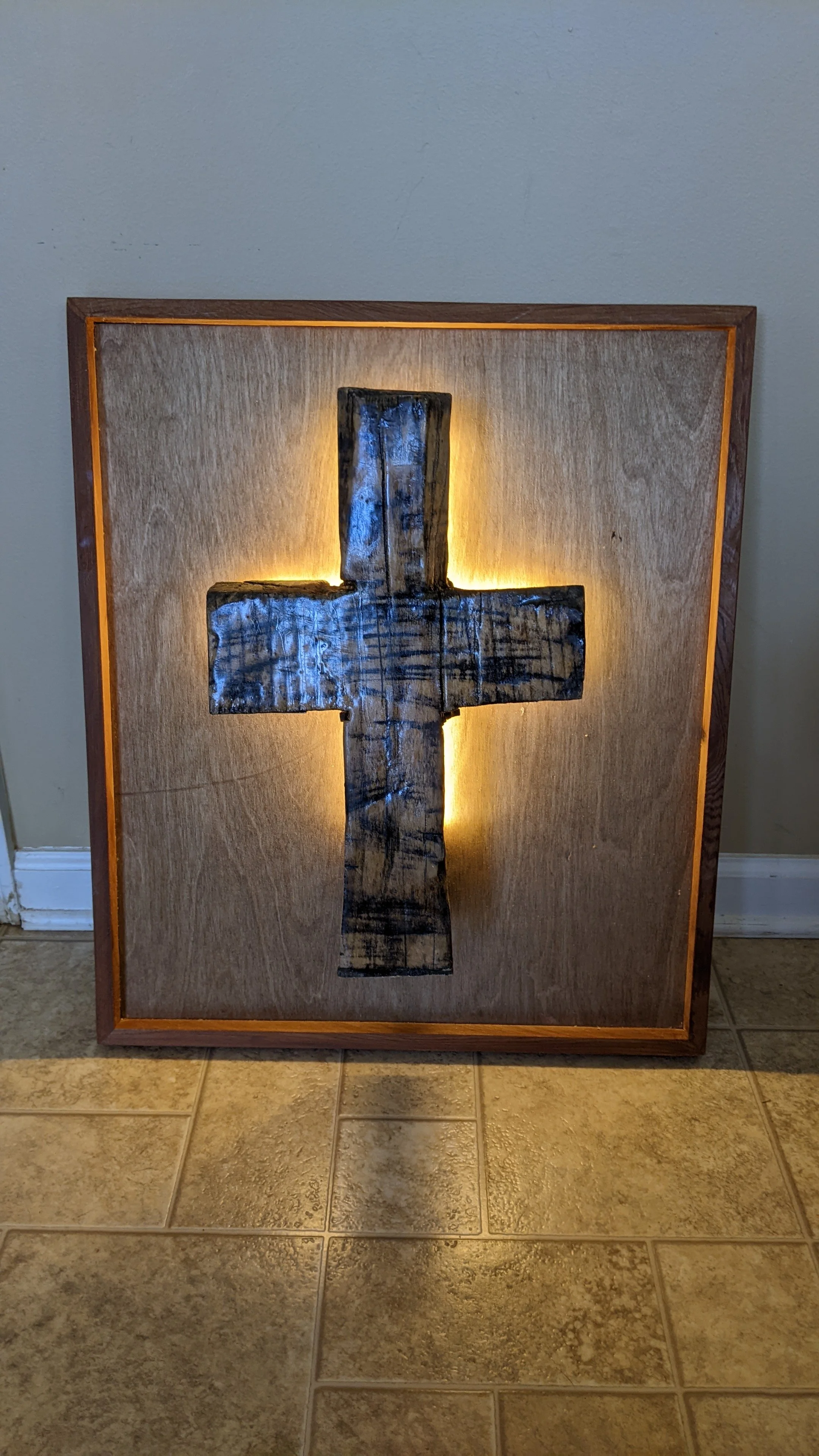 A wooden framed artwork featuring a cross with backlighting, illuminated from behind, standing on a tiled floor against a plain wall.