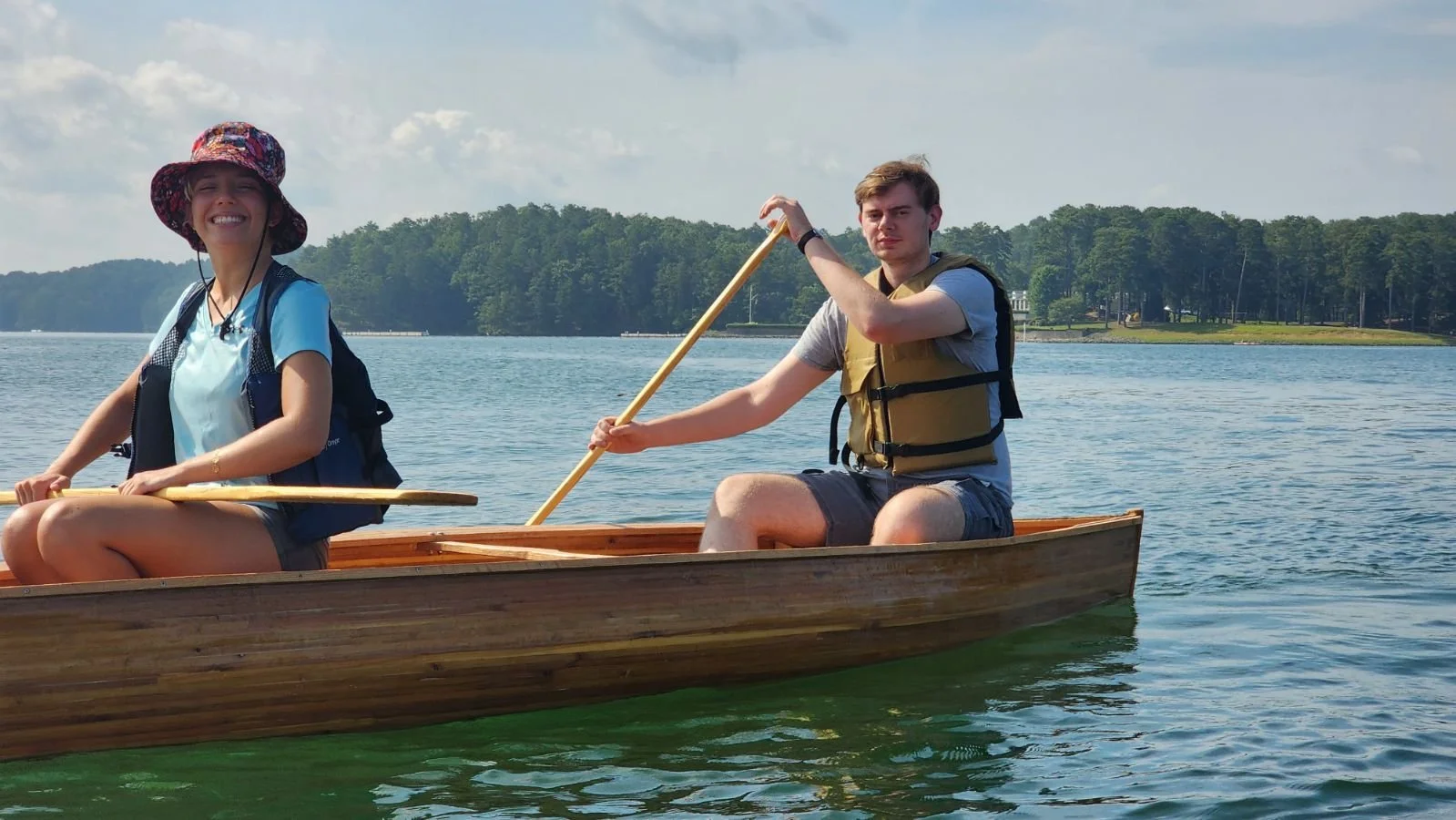 Two young people paddling a wooden canoe on a lake, with a tree-lined shore and partly cloudy sky in the background.