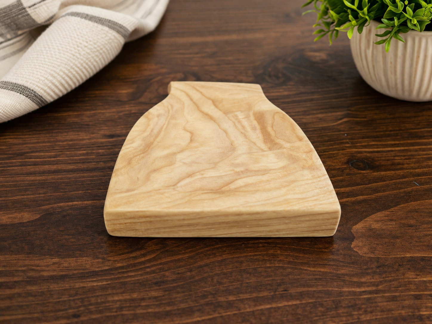 Wooden cutting board on a dark wood table with a green plant in a white pot and a white cloth with black stripes in the background.