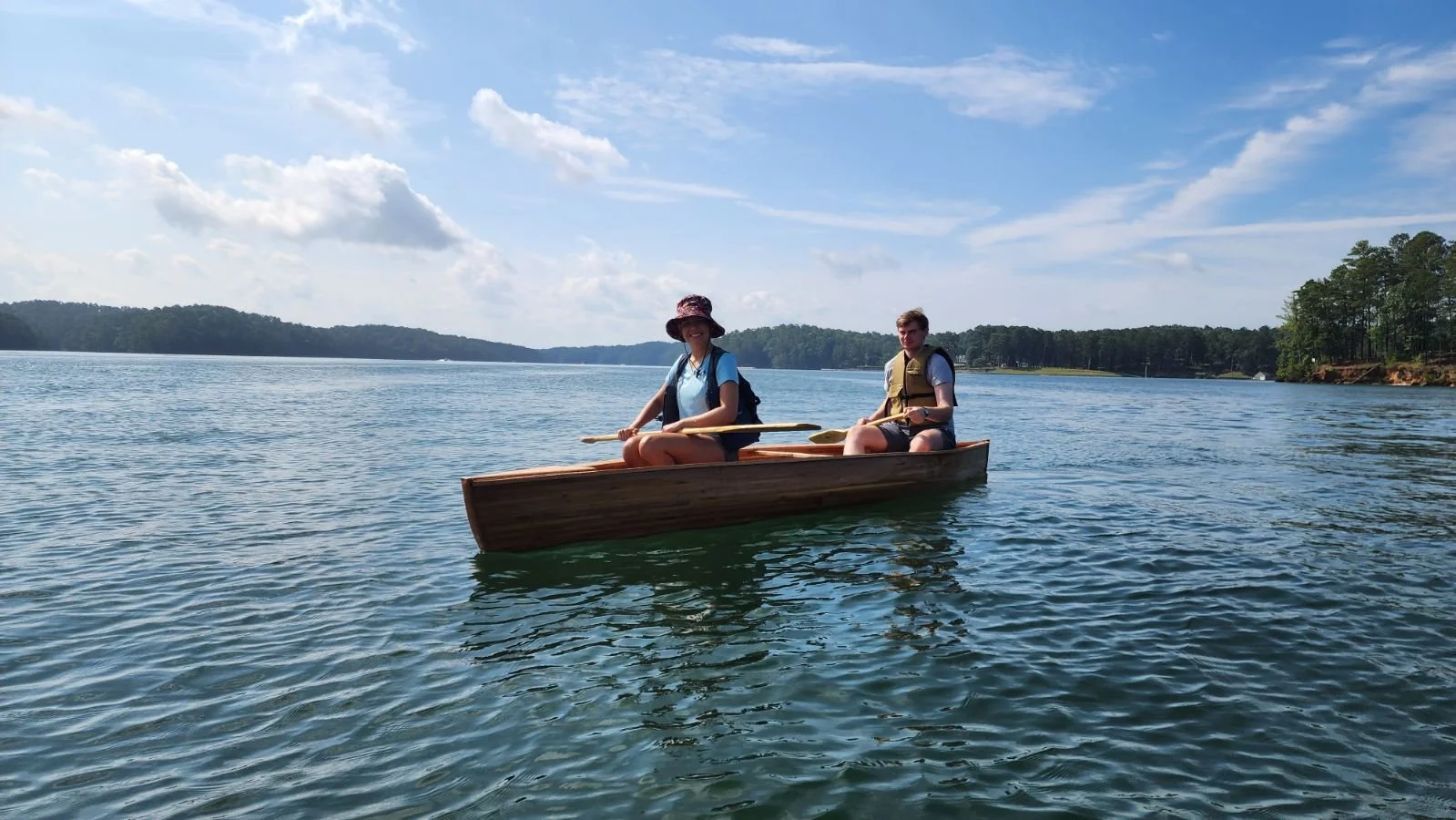 Two people in a canoe paddling on a calm lake under a partly cloudy sky, with distant trees on the shoreline.