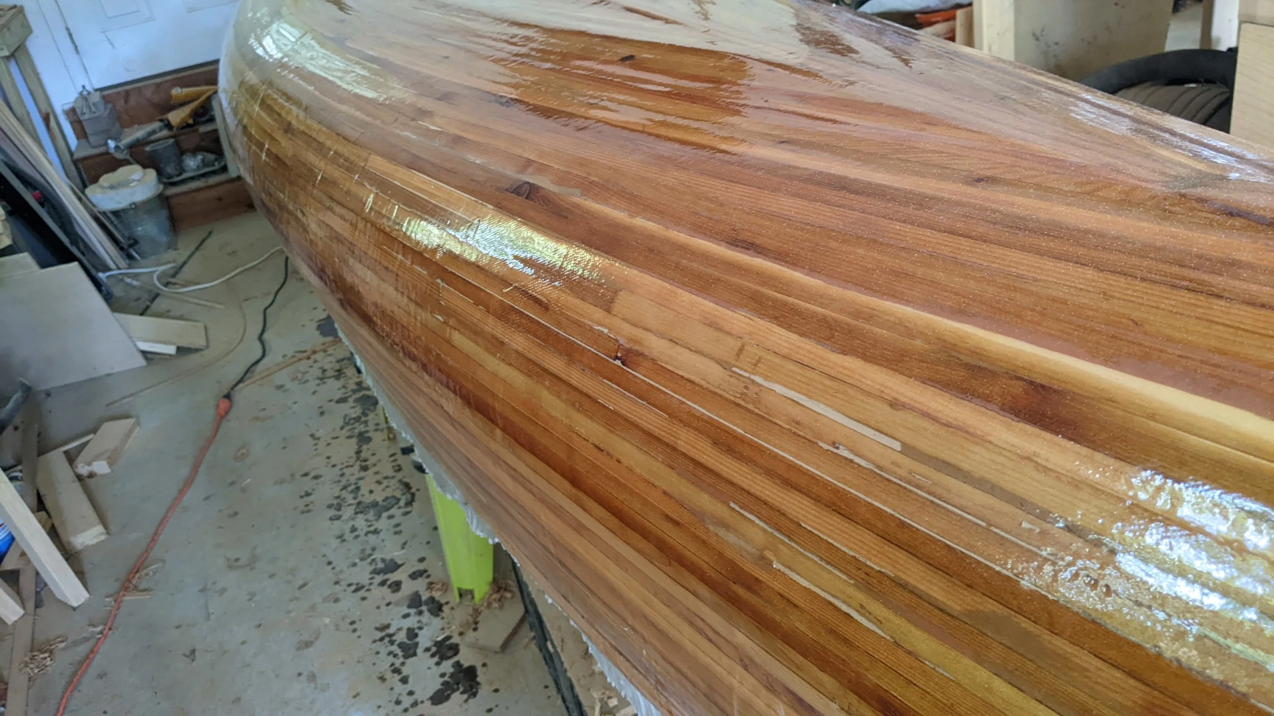 A wooden boat hull in a woodworking shop, wet and polished, sitting on a workbench.