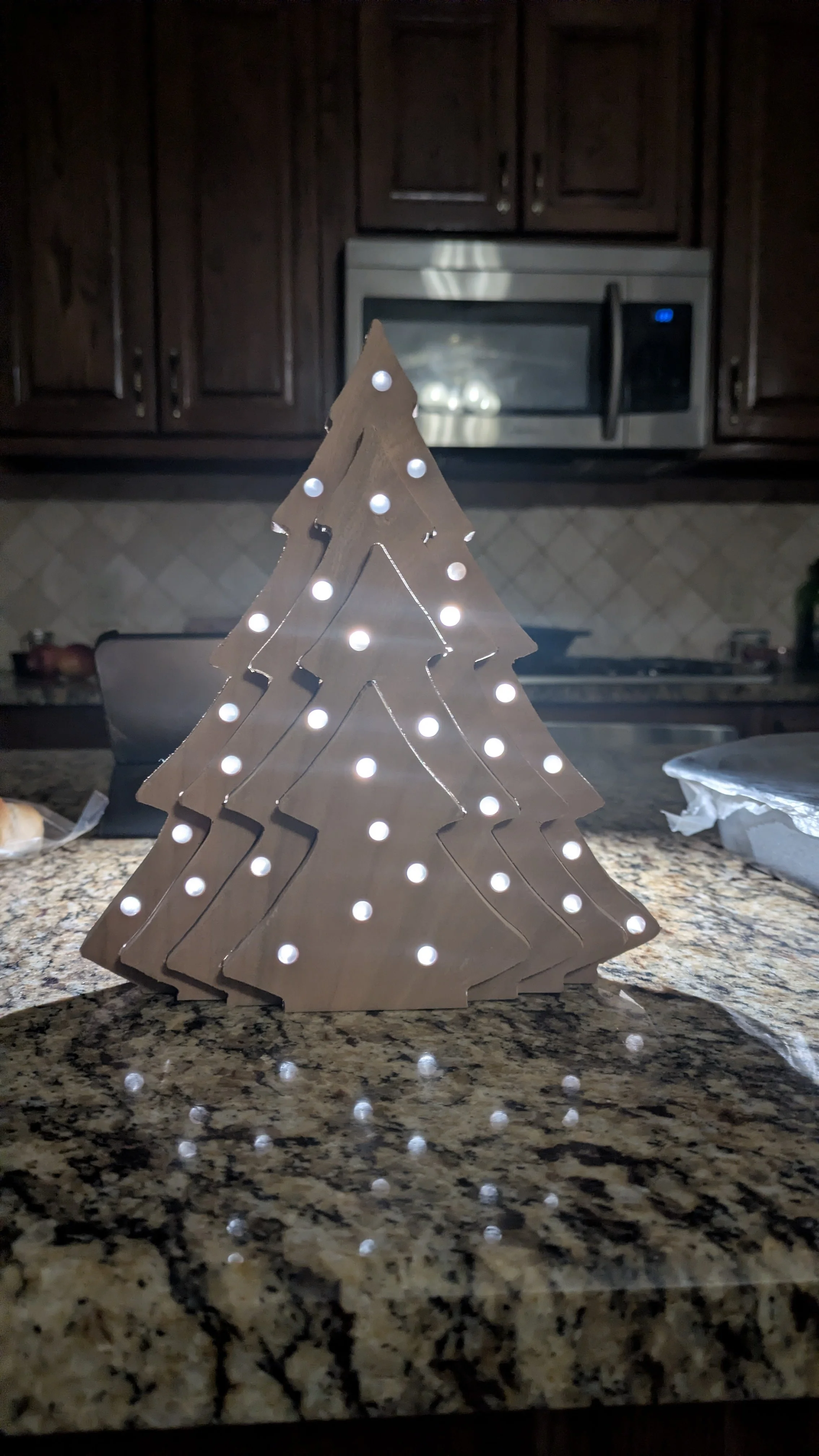 A decorative wooden Christmas tree with small white lights, placed on a granite countertop in a kitchen.