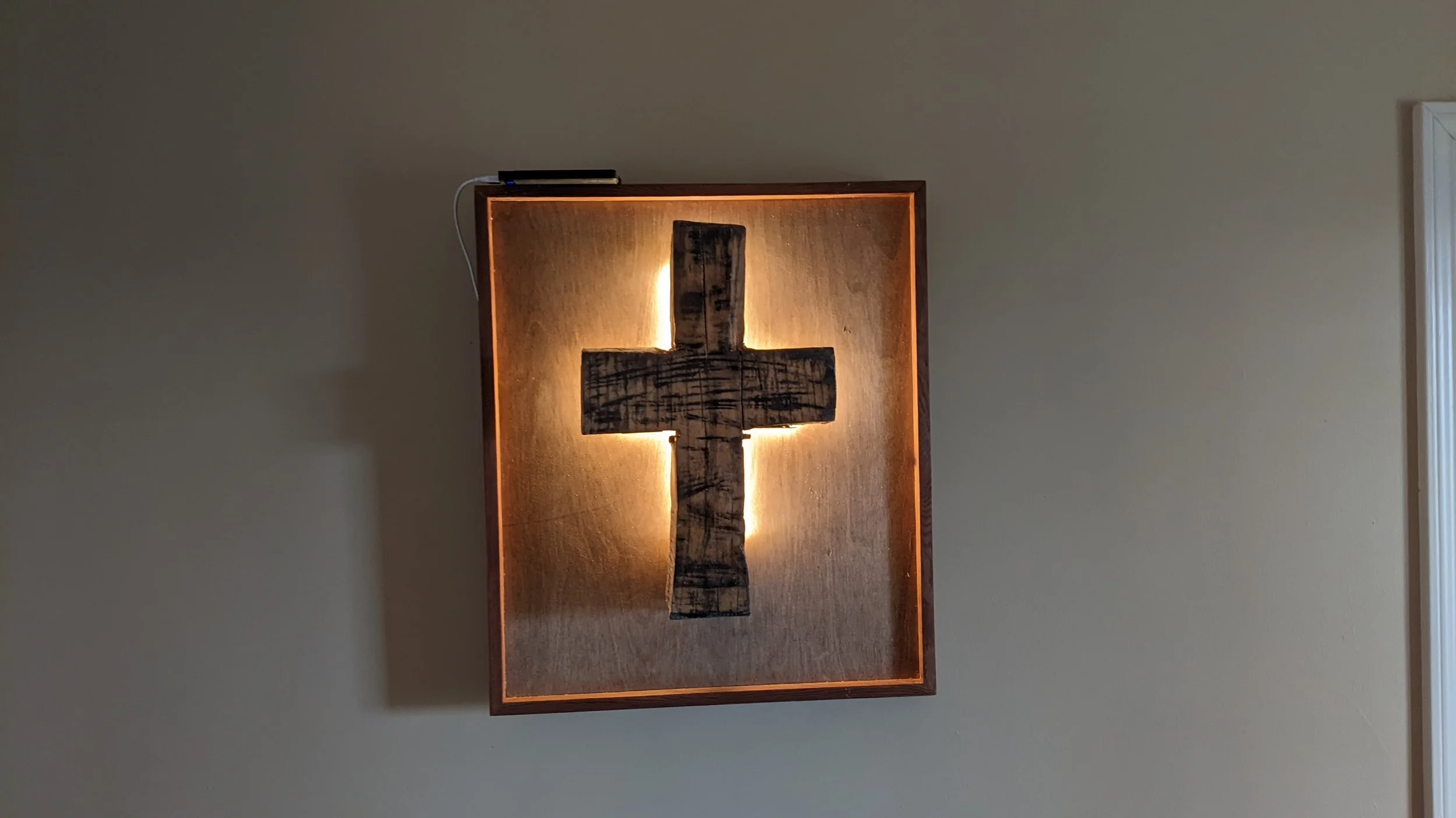 Wooden cross mounted on a wall with backlighting, in a wooden frame, and a black item connected by a white cord resting on top.