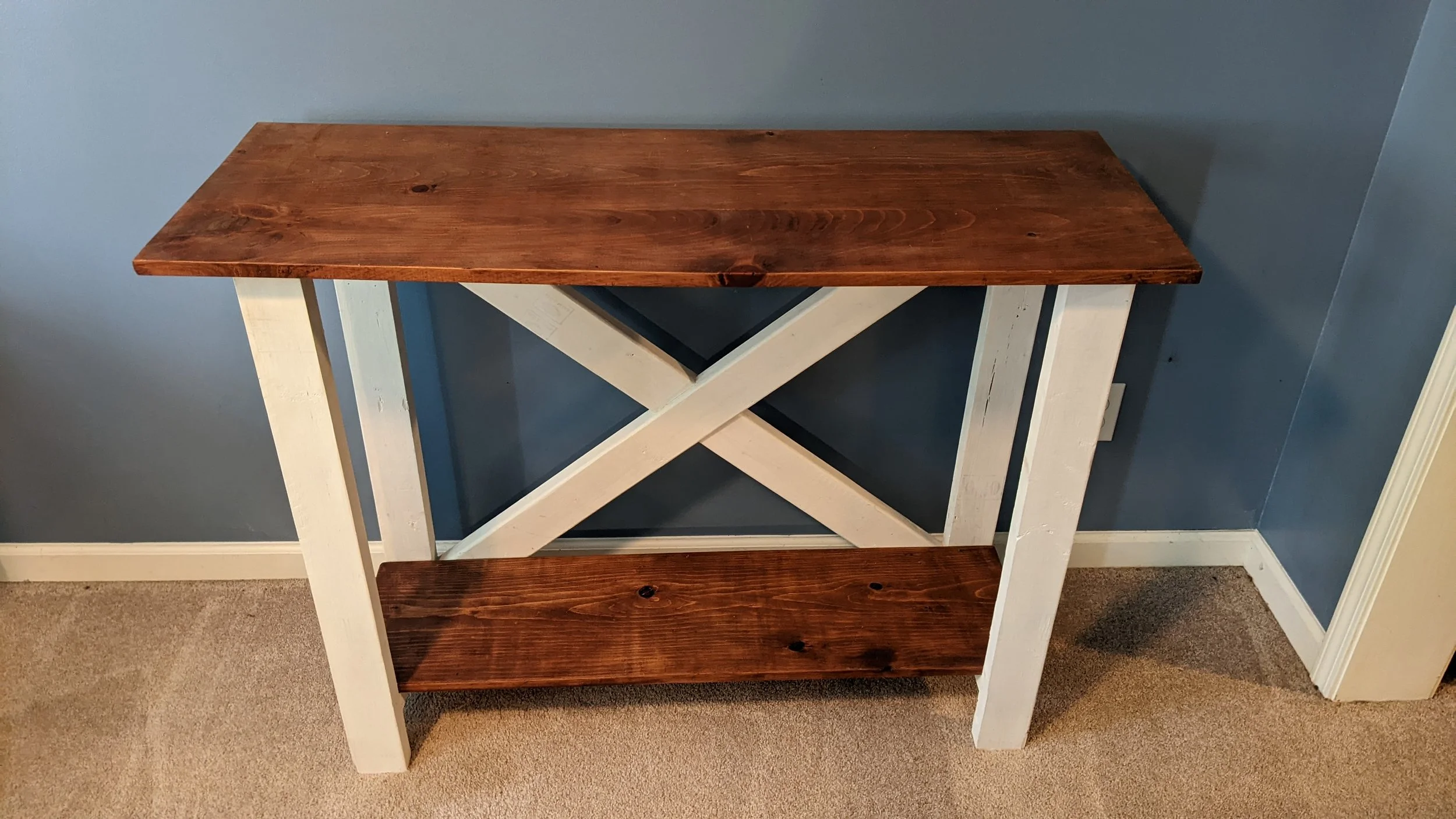 A wooden and white painted table or stand with two shelves, positioned against a blue wall near a carpeted floor.