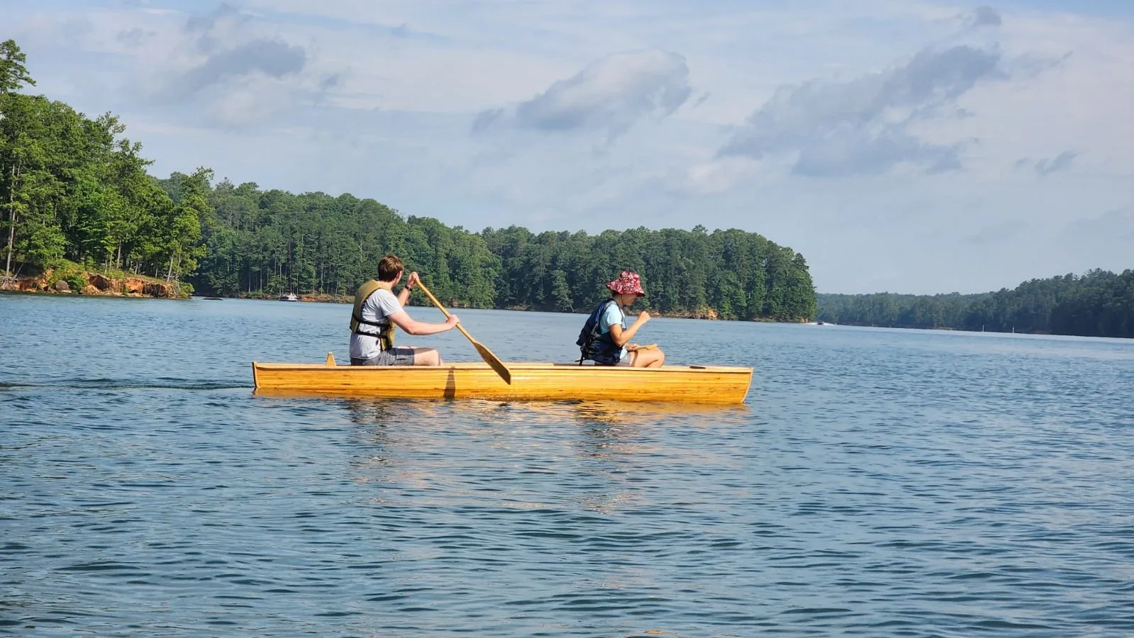 Two people kayaking on a river, surrounded by trees under a partly cloudy sky.
