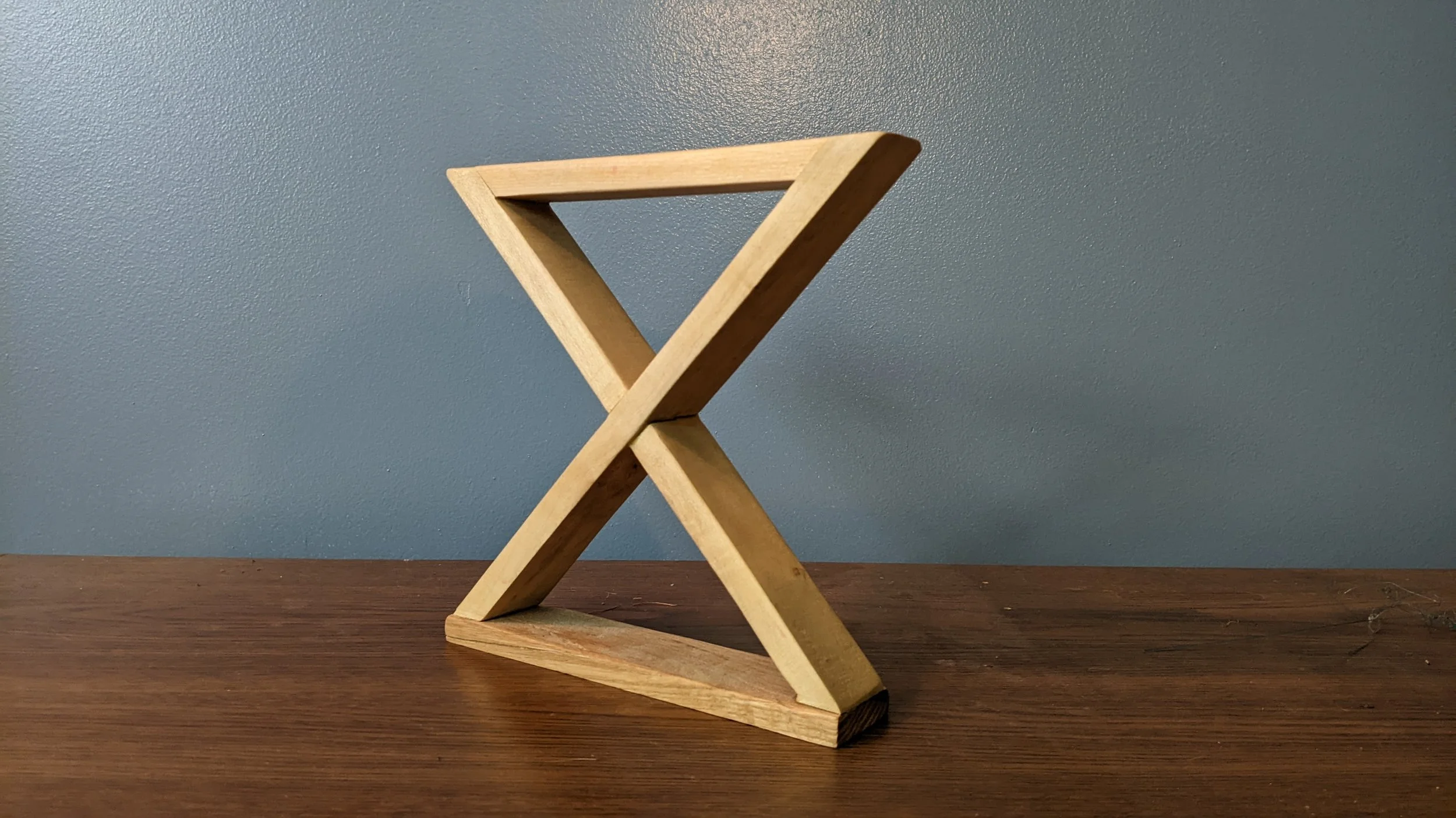 A small wooden folding stool with crossed legs on a wooden surface against a blue textured wall.