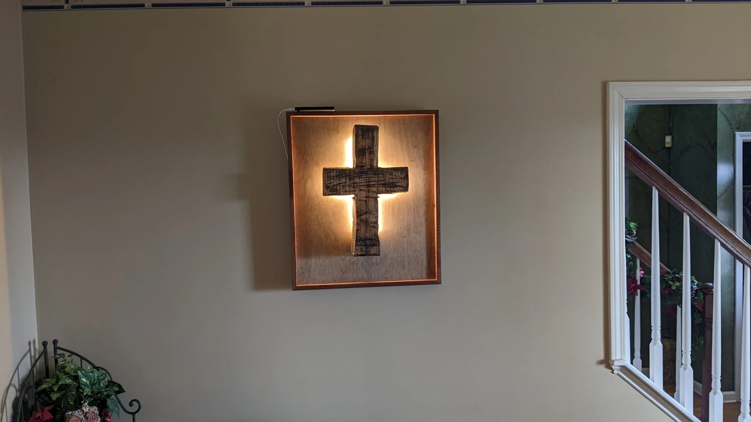 Wall hanging sculpture of a cross illuminated with backlighting, mounted on a beige wall near a staircase with white railing and greenery visible through a window.