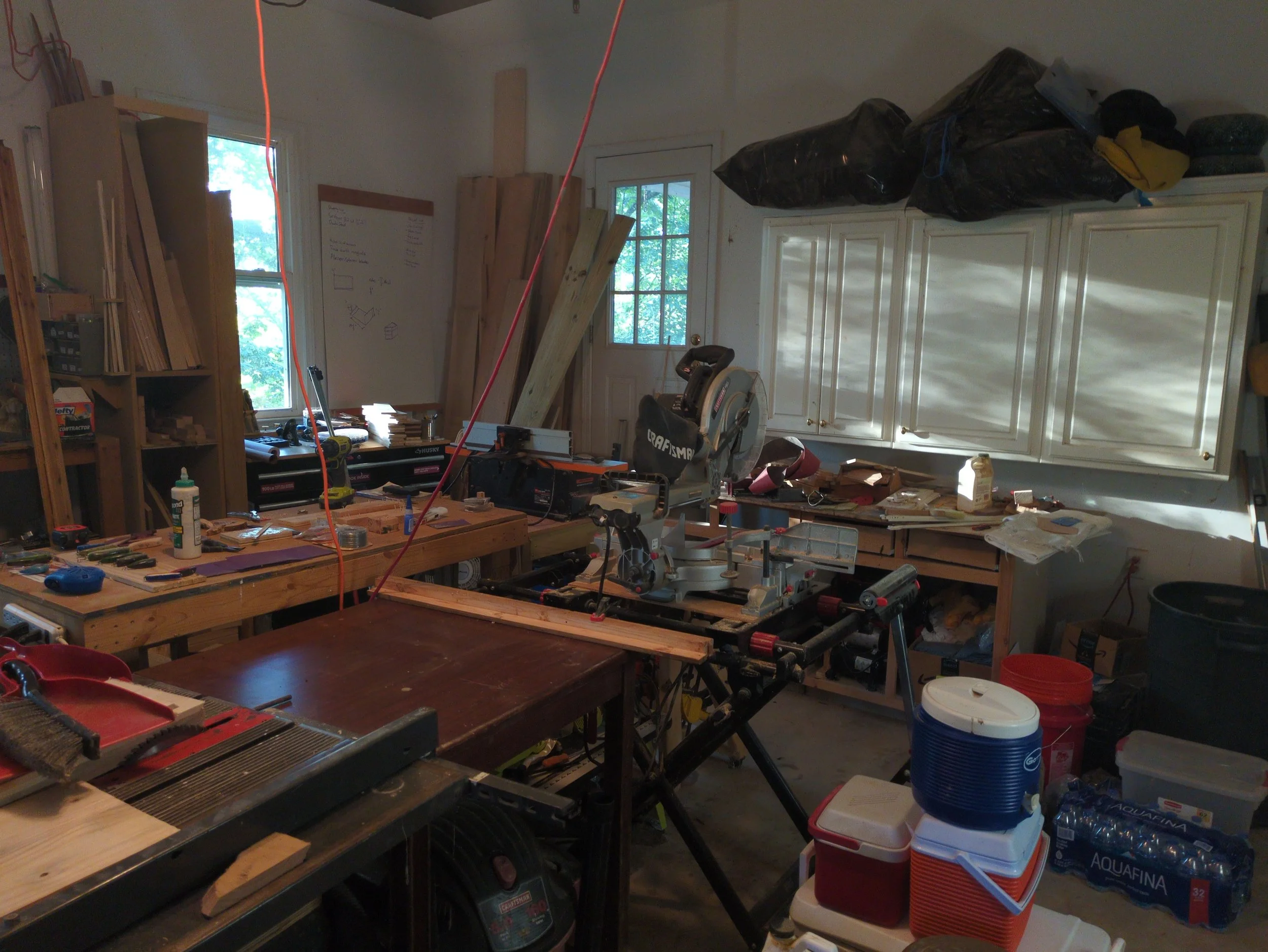A cluttered workshop or garage space with woodworking tools, wooden planks, storage shelves, and various supplies such as water bottles, buckets, and a miter saw.