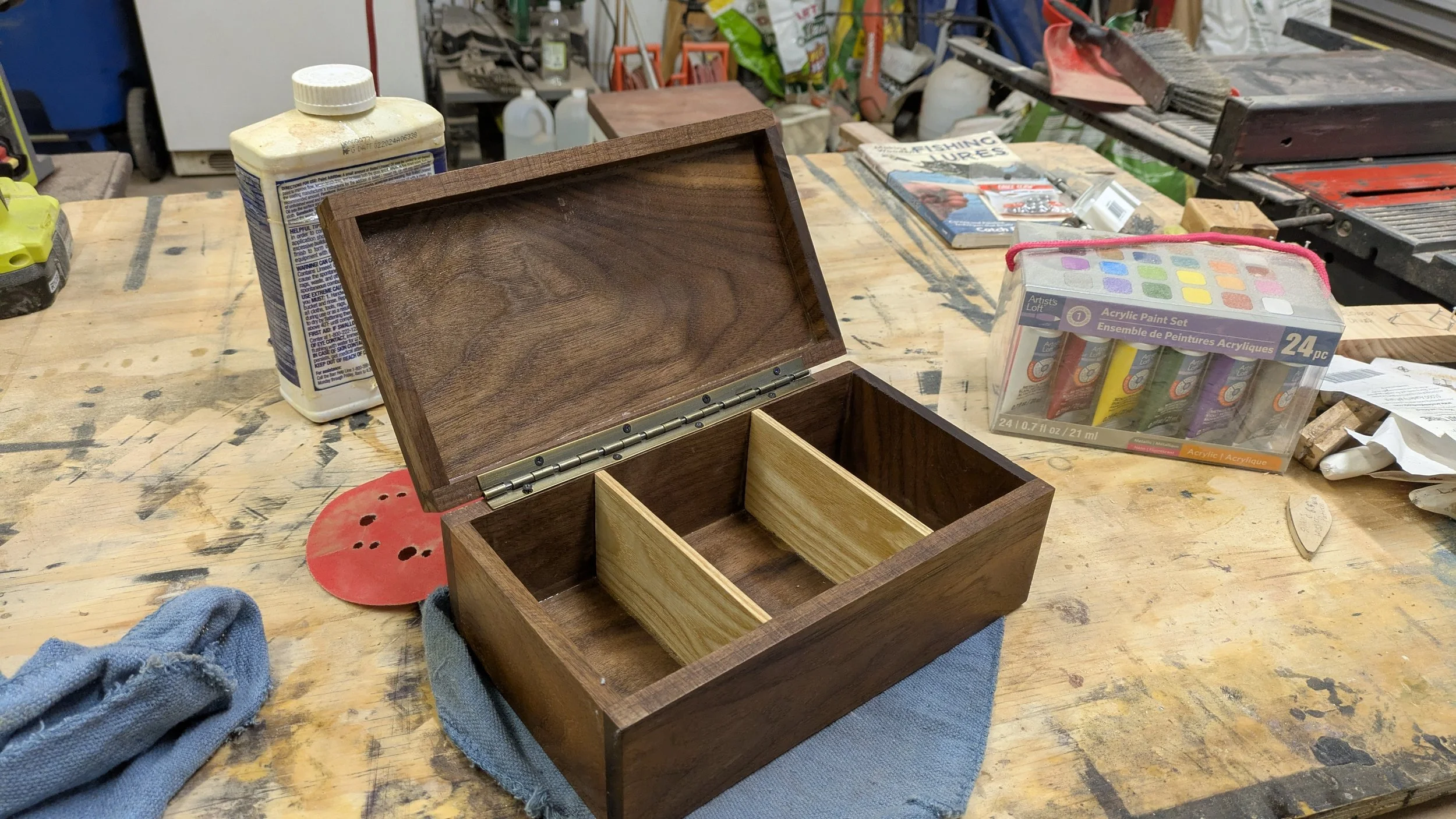 Open wooden jewelry box on a workbench in a woodworking shop, with crafting supplies and tools in the background.