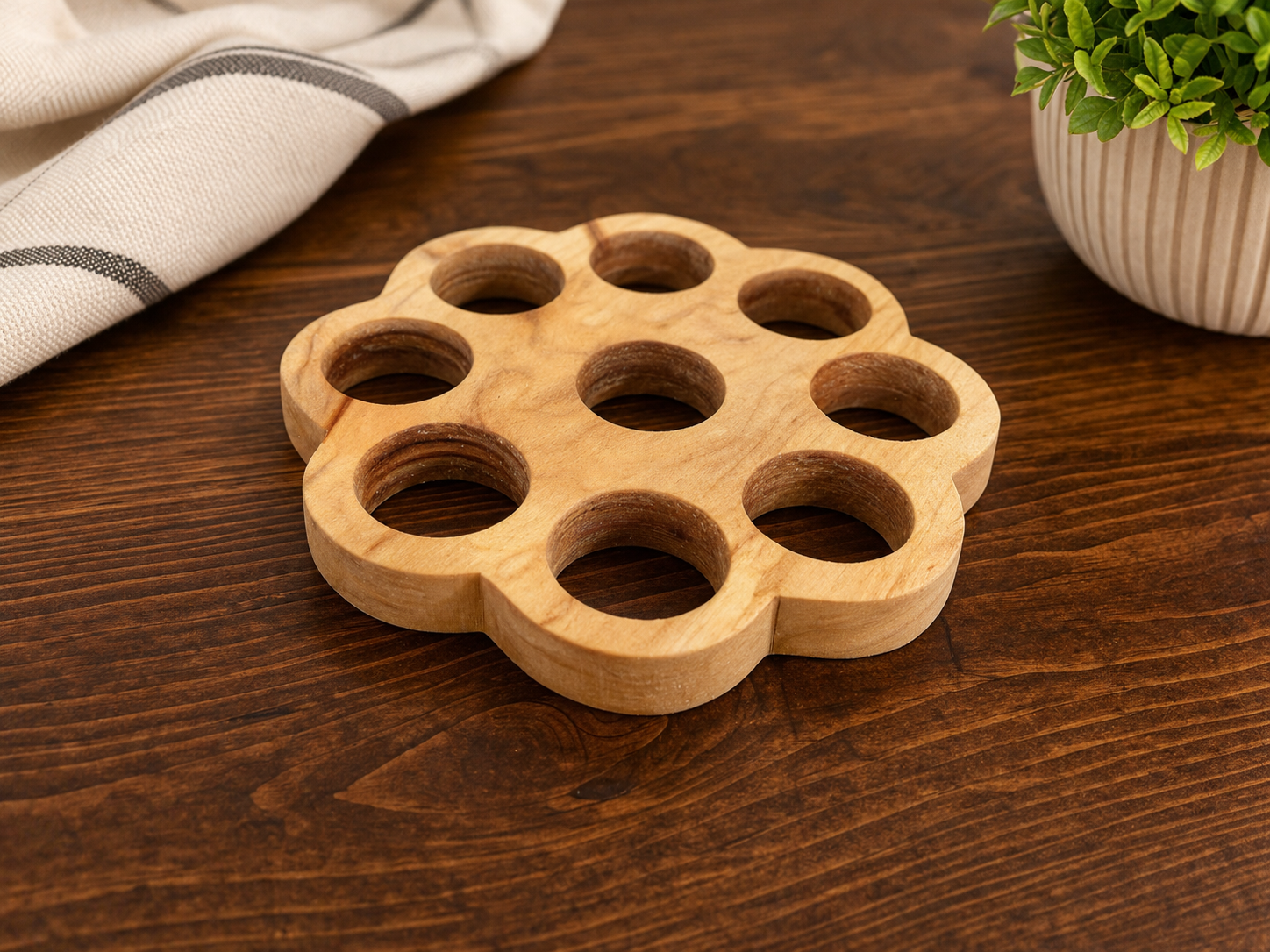 A wooden circular object with multiple large holes on a dark wooden table, next to a white and gray striped cloth and a potted green plant.