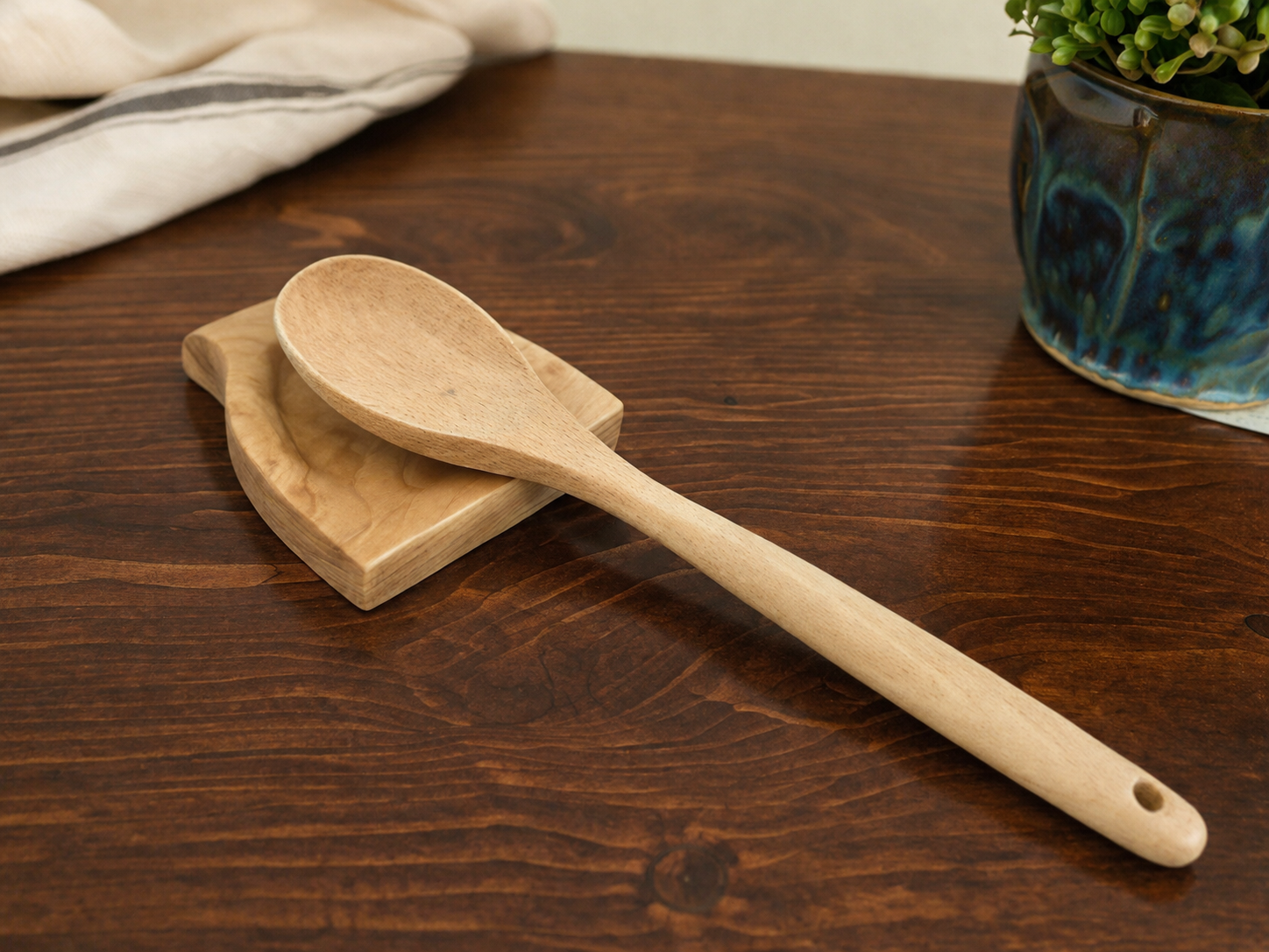 A wooden spoon resting on a wooden cutting board on a dark wood table, with a ceramic pot with green plants in the background and a fabric cloth in the upper left corner.