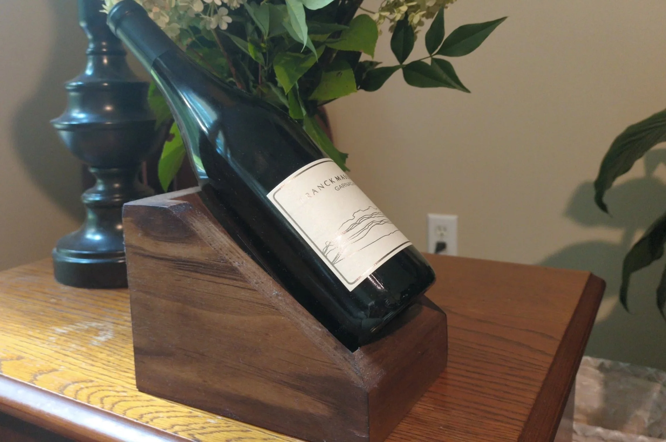 A wine bottle with a minimalist label, resting on a wooden stand on a wooden table. In the background, there is a dark metal candlestick holder and some green leafy plants with white flowers.