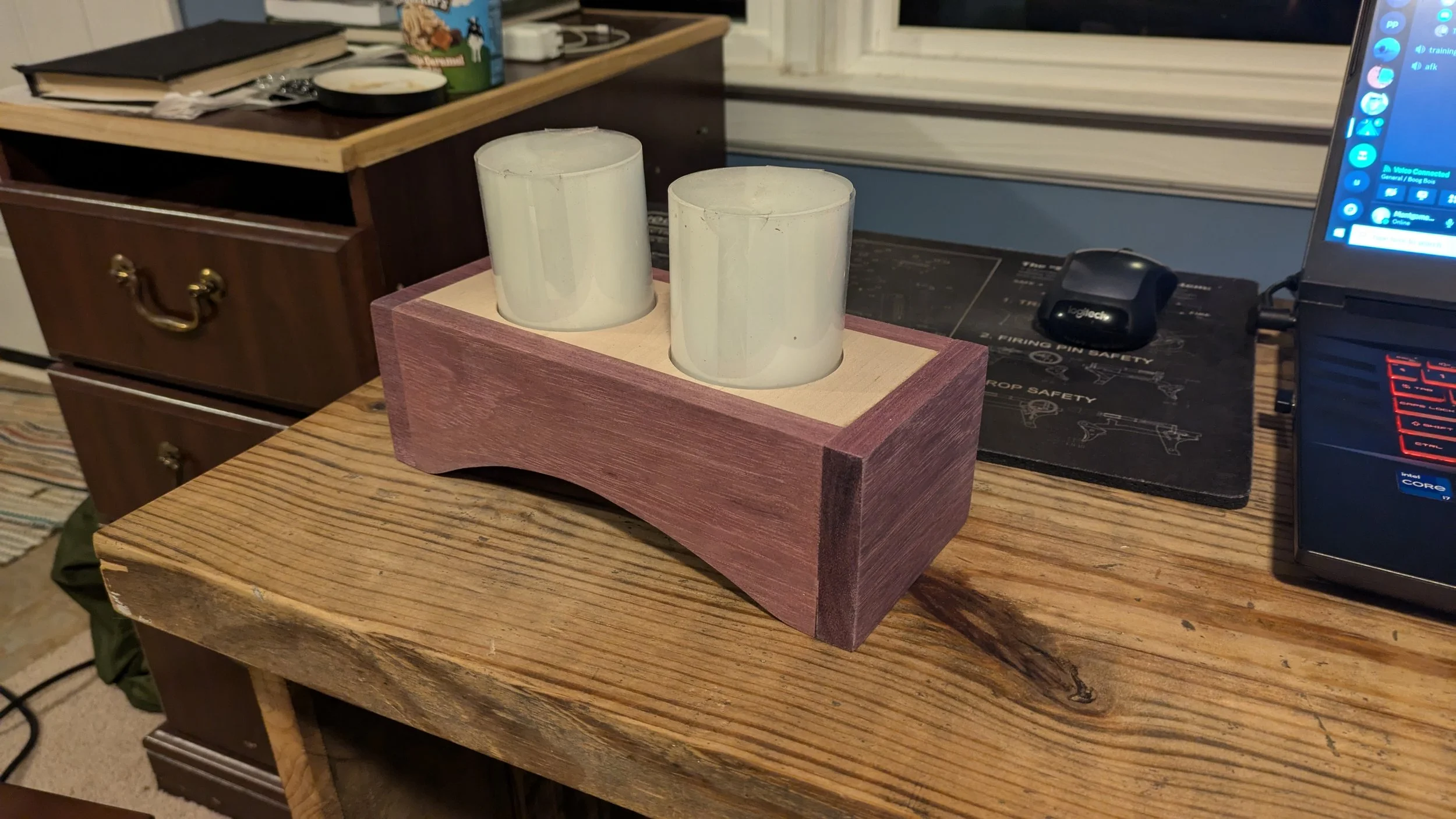 A wooden table holding a rectangular wooden holder with two white candles. In the background, there are various items including a black book, a cereal box, a bowl, a computer mouse, and a laptop on a desk.